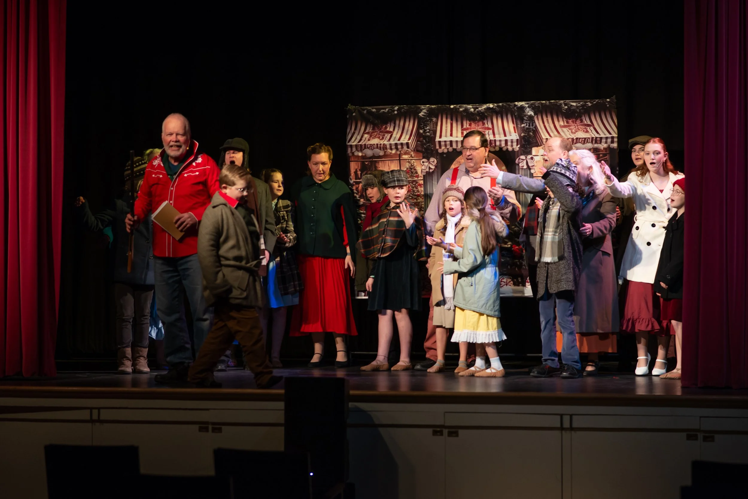 People acting in a holiday play on stage with Christmas backdrop, including children and adults dressed in costumes, some holding scripts or gesturing.