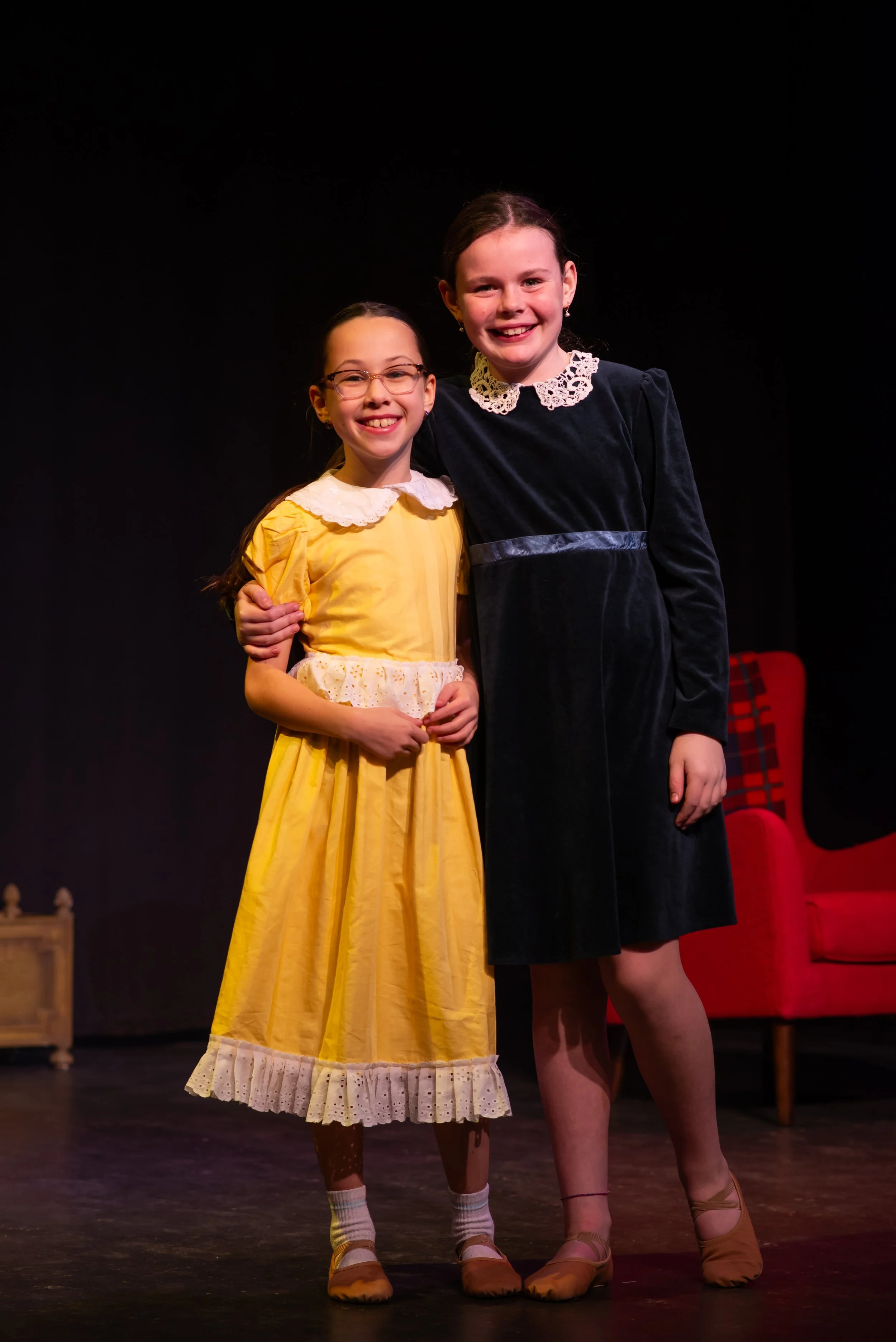 Two young girls standing on stage, smiling, posing for a photo. One girl is wearing a yellow dress with a white lace collar and glasses, while the other girl is dressed in a dark velvet dress with a lace collar. There are red chairs and a small table