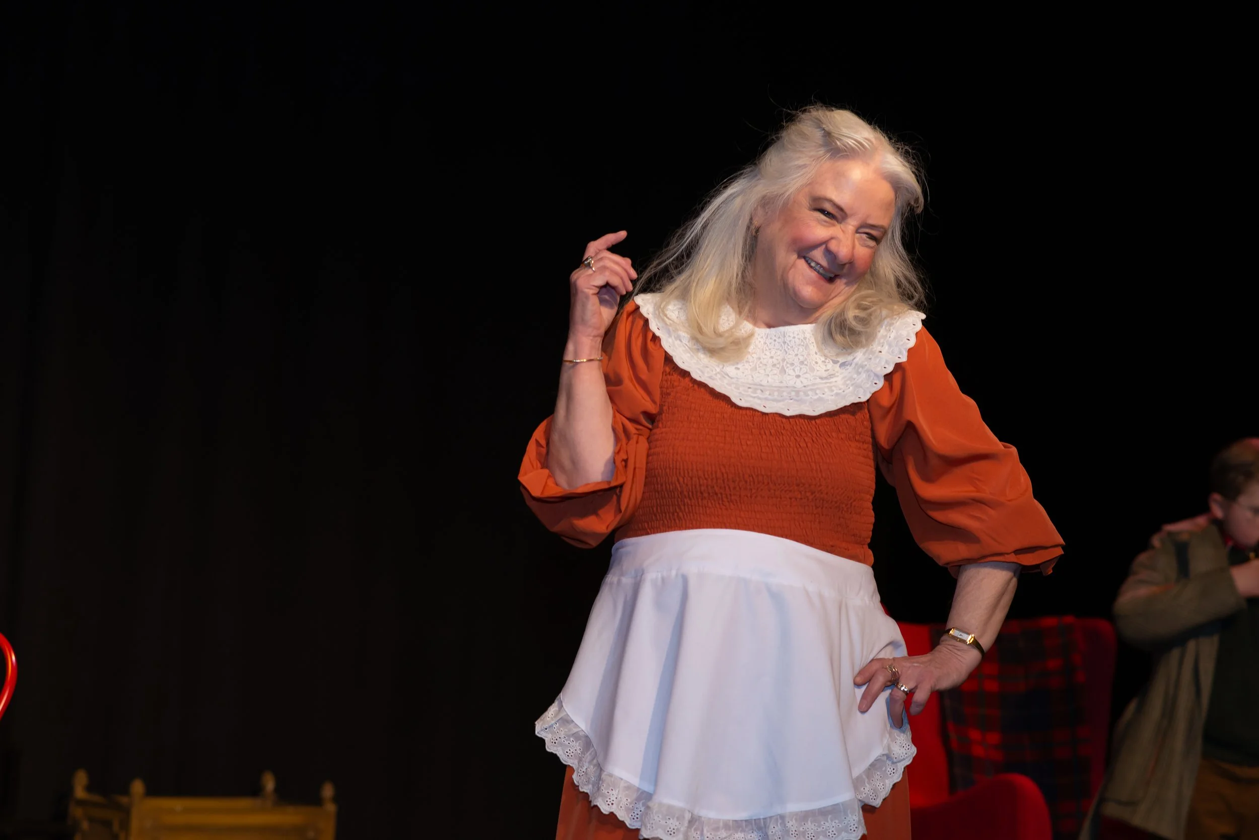 A woman with gray hair dressed in an orange top with a white lace collar and a white skirt, smiling and gesturing with her right hand, in a dark room with a black background.