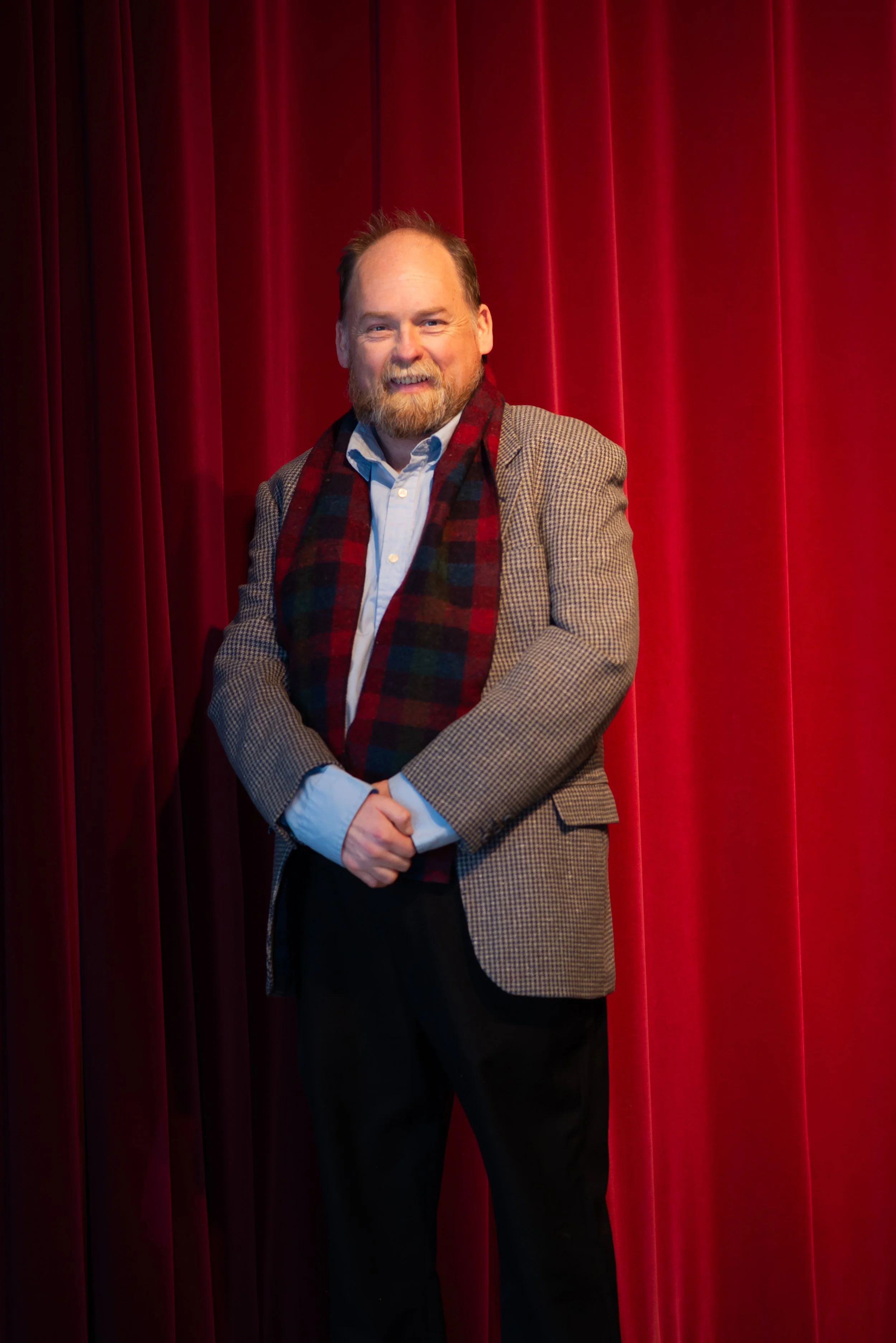 A man with a beard dressed in a plaid scarf, blazer, and light blue shirt standing in front of red theater curtains.