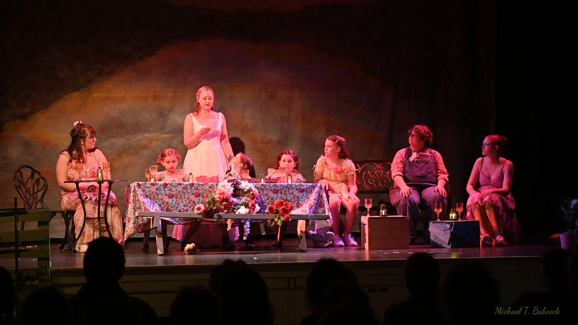 A theatrical stage scene with seven children and one adult woman, seated or standing around a table decorated with a floral cloth and flowers. The woman is reading from a paper, and the children display attentive and curious expressions. The scene su