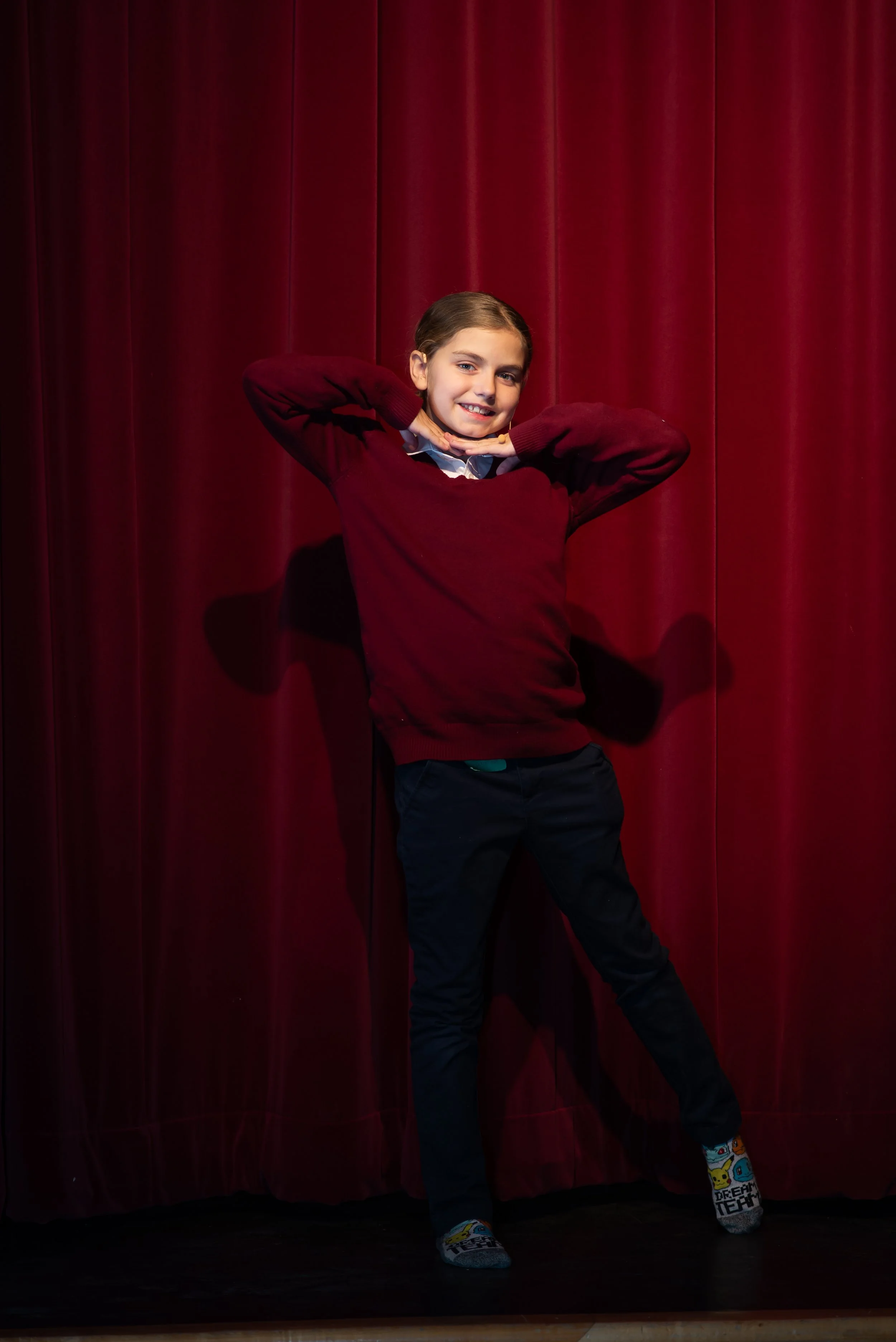 A smiling girl in a maroon sweater posing with arms behind head in front of red curtains