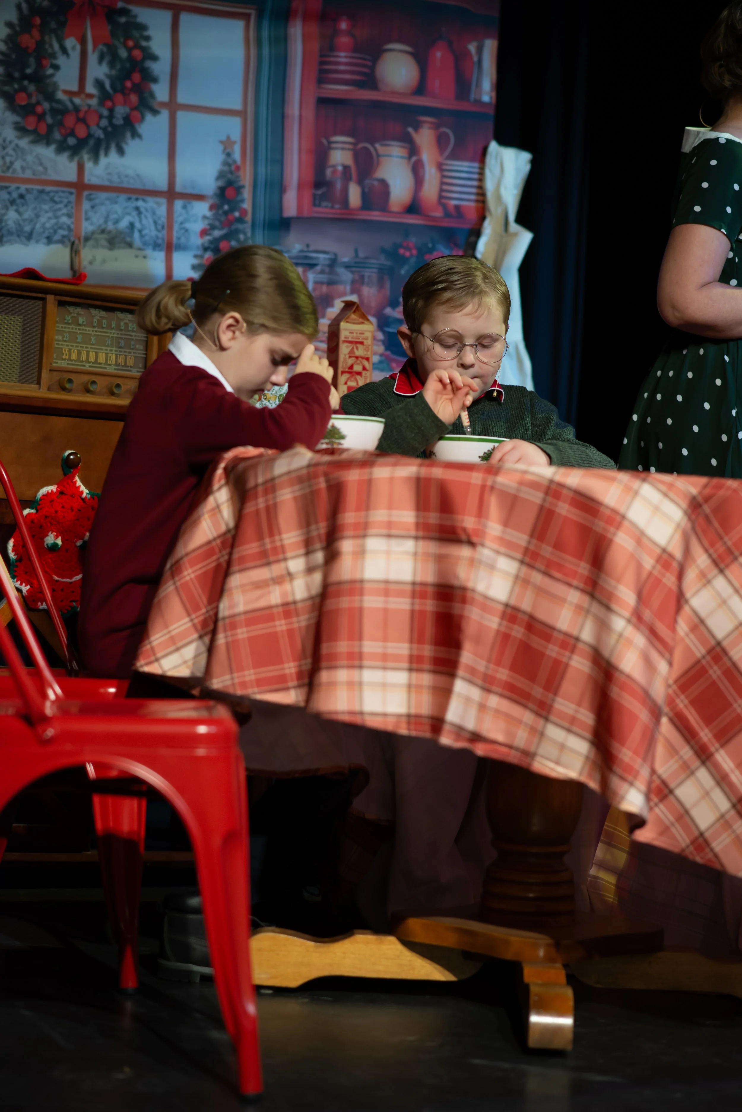 Two children sitting at a table with a red checkered tablecloth, enjoying a meal, with holiday decorations in the background.
