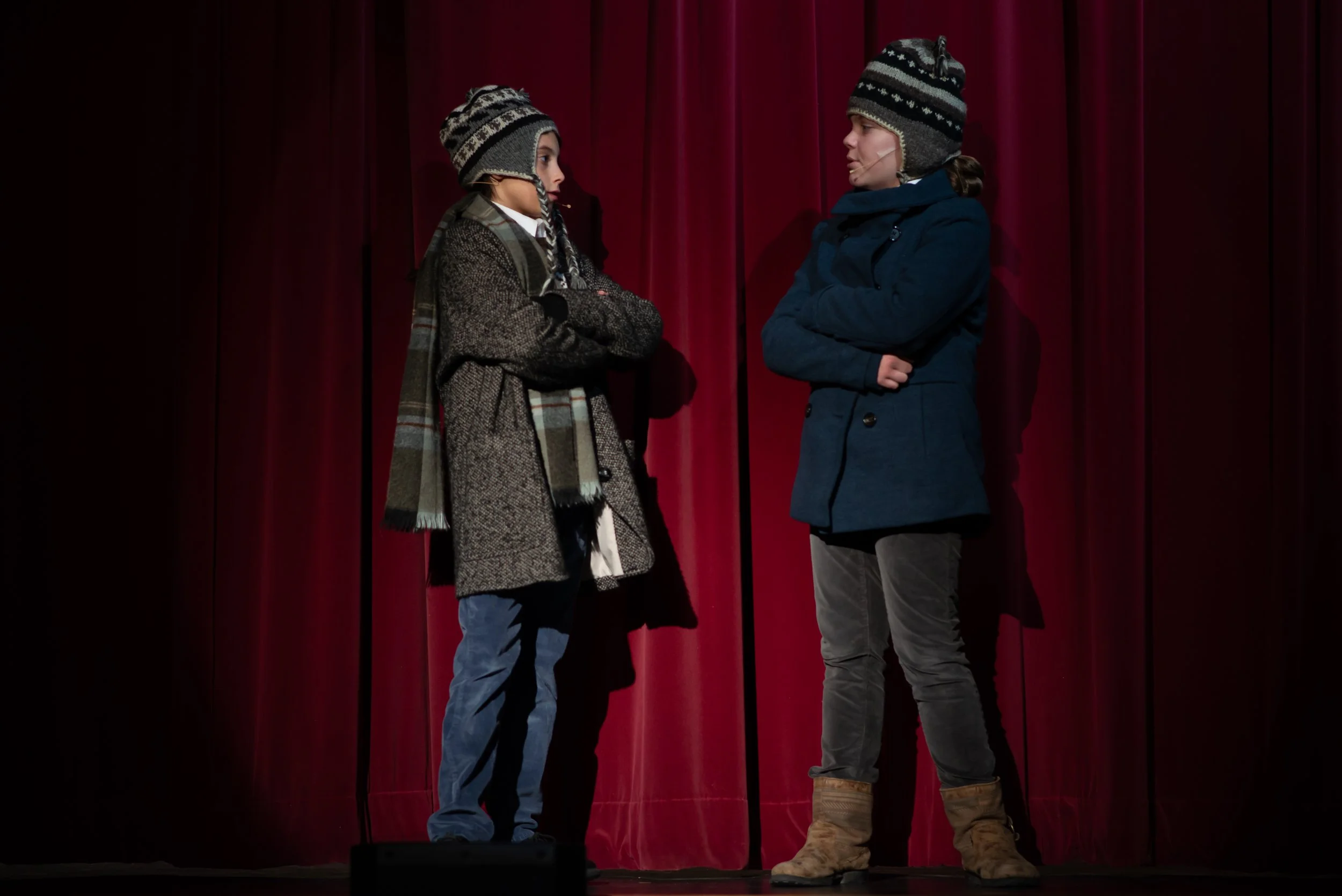 Two children stand on a stage in front of a red velvet curtain, facing each other with arms crossed, wearing winter clothes and knit hats.