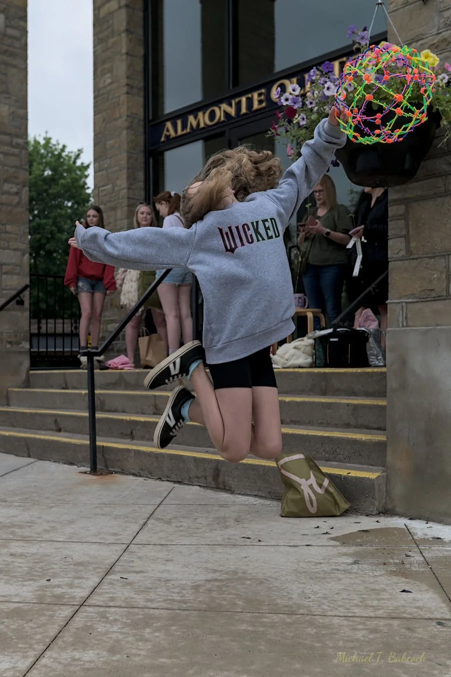 Girl jumping in front of a building labeled 'Almonte' with people standing on the steps behind her. She is wearing a gray hoodie with 'WICKED' on the back, black shorts, and sneakers, holding a colorful balloon sculpture, and has a bag on the ground.
