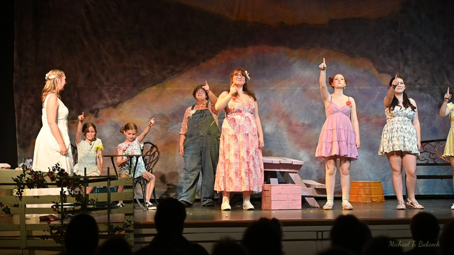Group of young girls and women on stage during a performance; some are standing, some are sitting, and they are dressed in colorful, casual dresses and overalls. The background is a painted backdrop resembling a natural scene, and there are props lik
