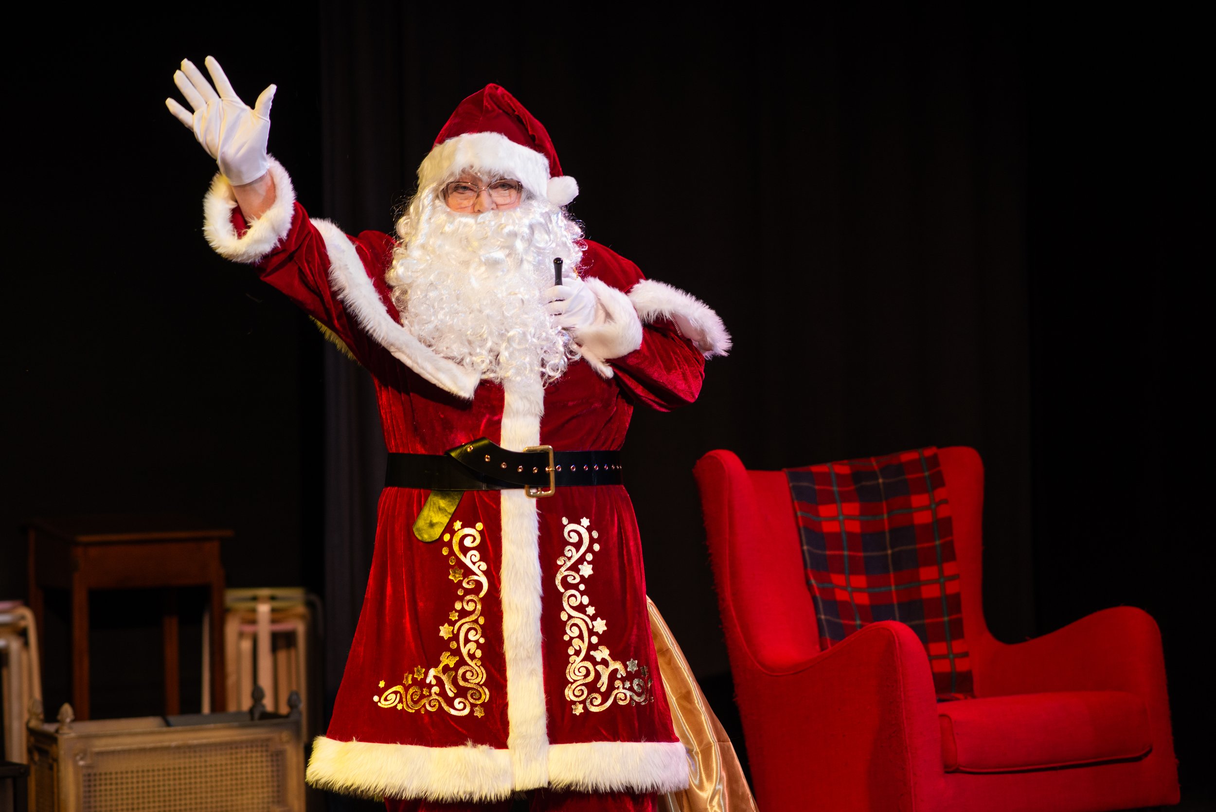 Santa Claus waving and smiling on stage in front of a black background, wearing a traditional red and white suit, glasses, and holding a microphone, with red velvet chairs and holiday decorations in the scene.