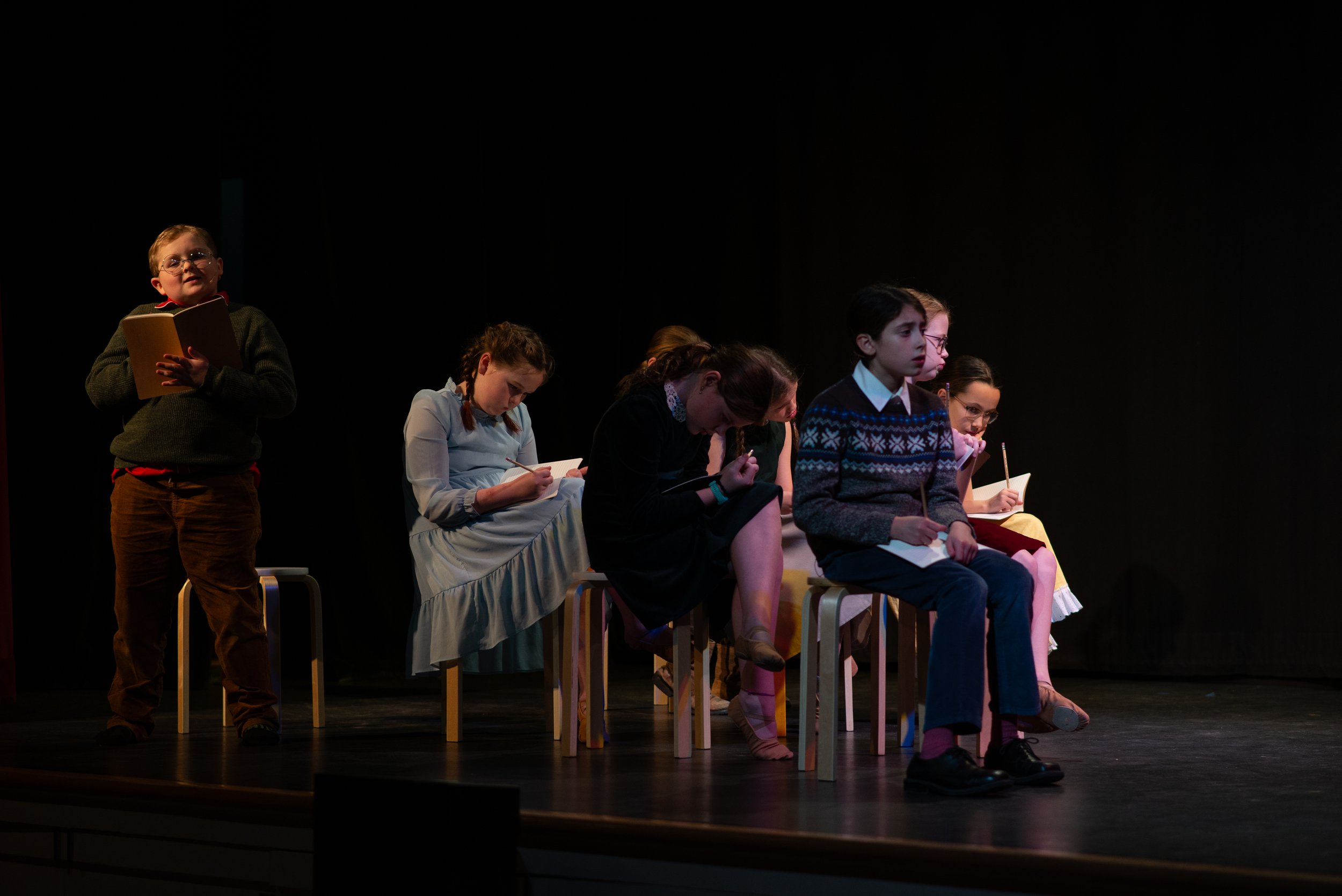 Children sitting on stage in a theatrical performance, with one boy standing and reading from a book while others are seated, some taking notes or holding notebooks, against a dark background.