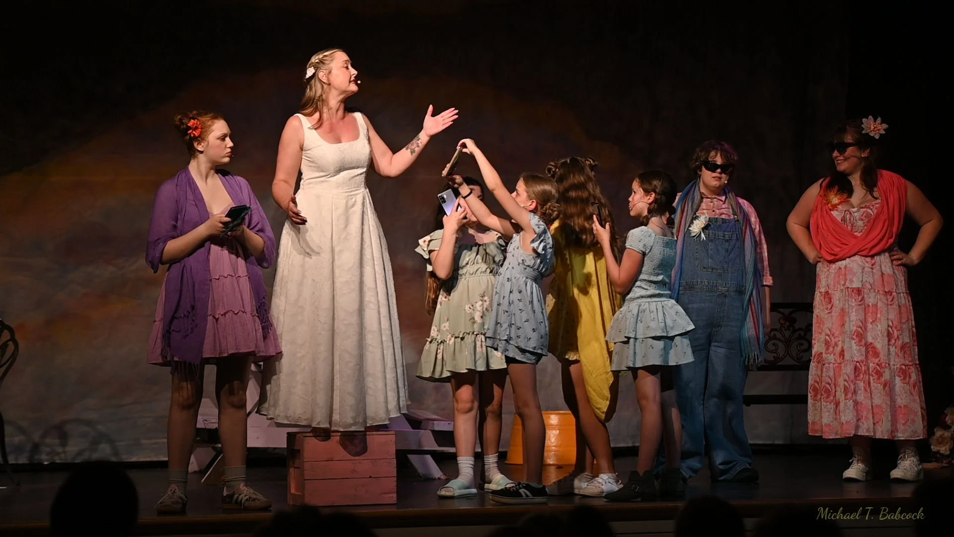 A theatrical scene with nine women on stage, some dressed casually and others in costumes. The woman in the center wears a white dress and is speaking to a group of young girls, who are reaching out to her. Other women, including one in sunglasses an