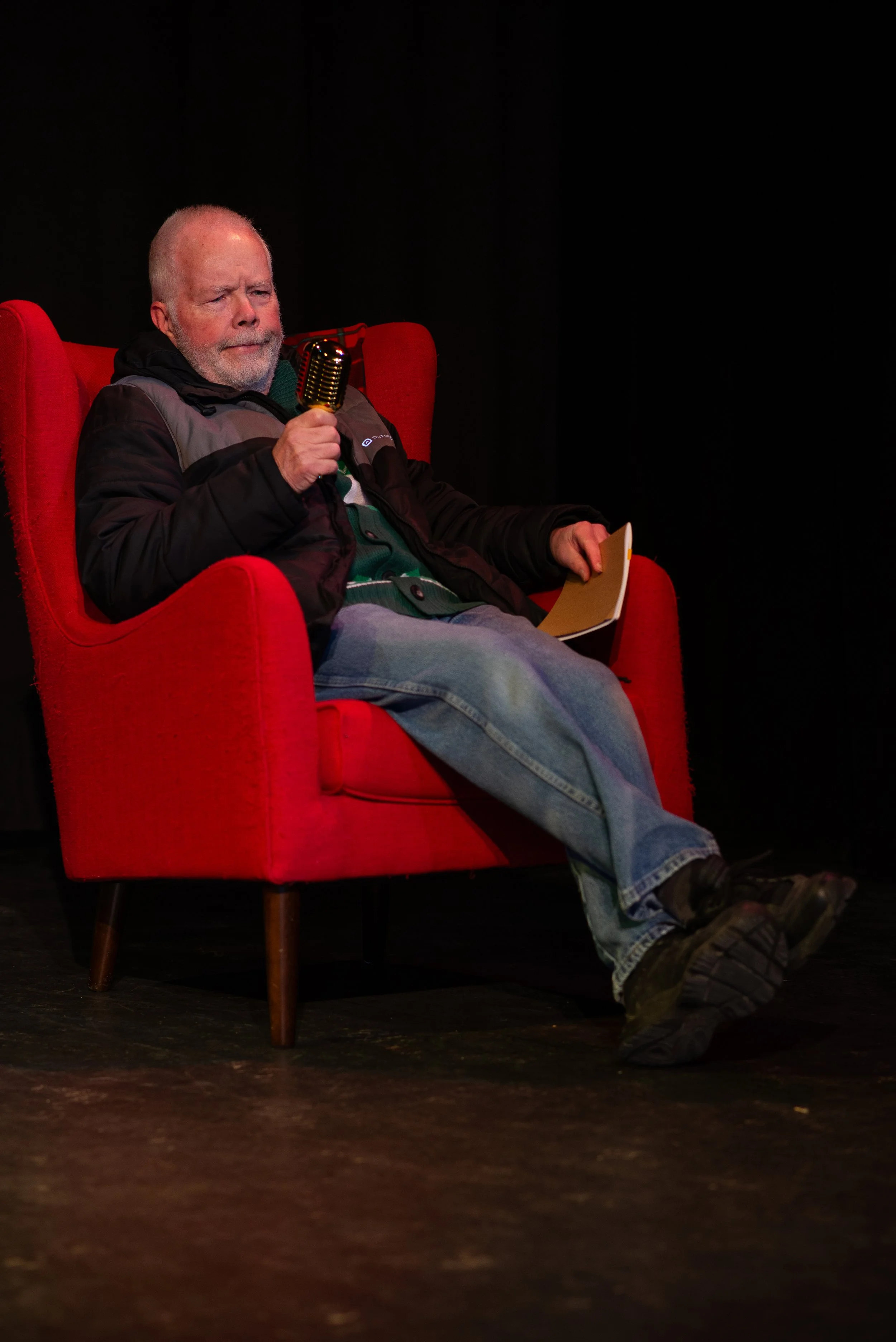Older man with a beard sitting in a red armchair on a dark stage holding a microphone and a notepad.