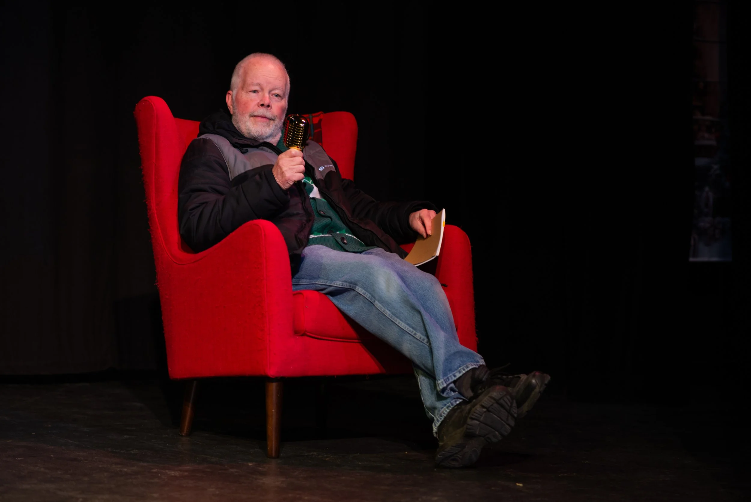 An older man with a beard sitting in a red armchair holding a vintage microphone, dark background, casual clothing