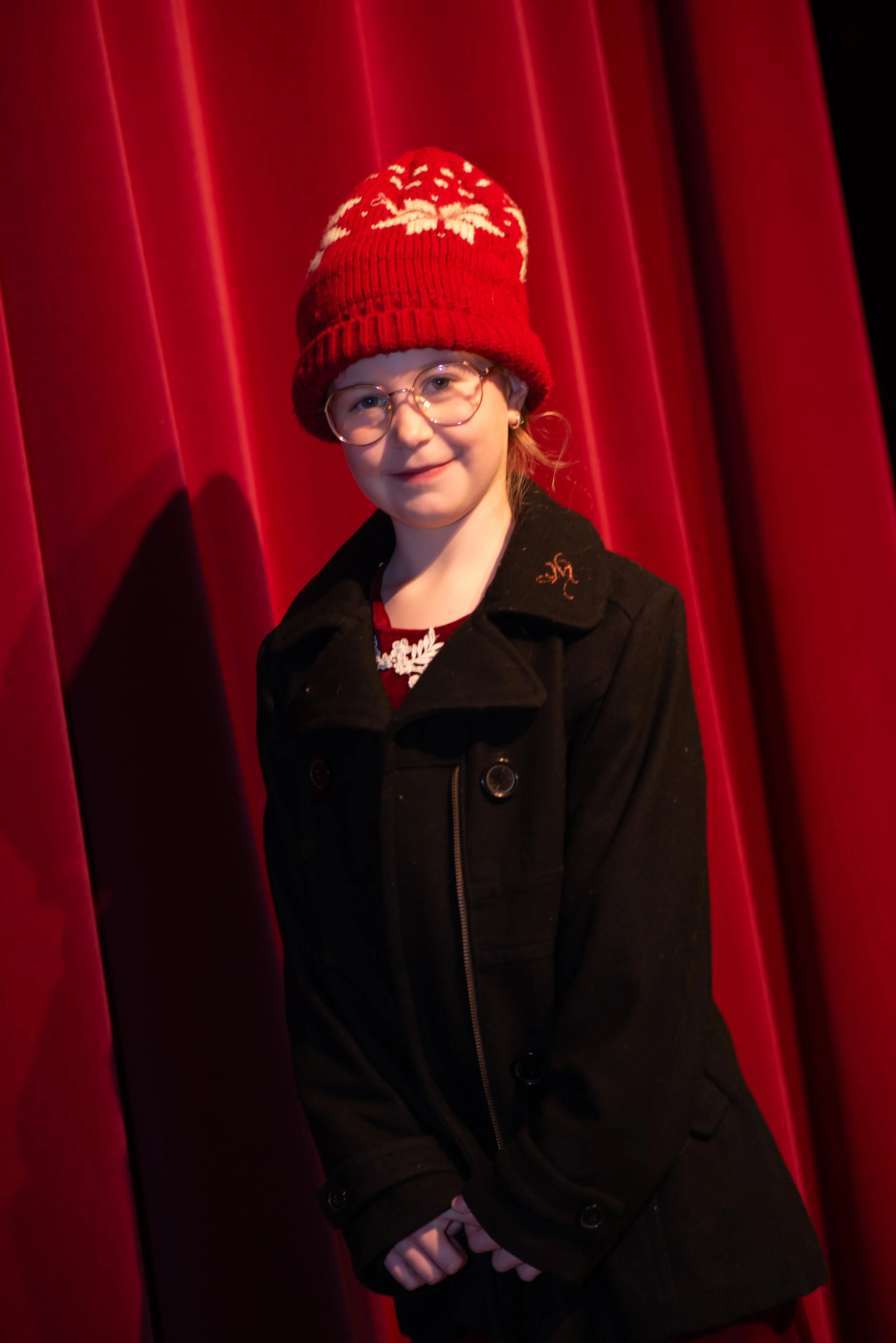 A young girl with glasses and a red knit hat with white patterns, standing in front of red stage curtains, wearing a black coat.