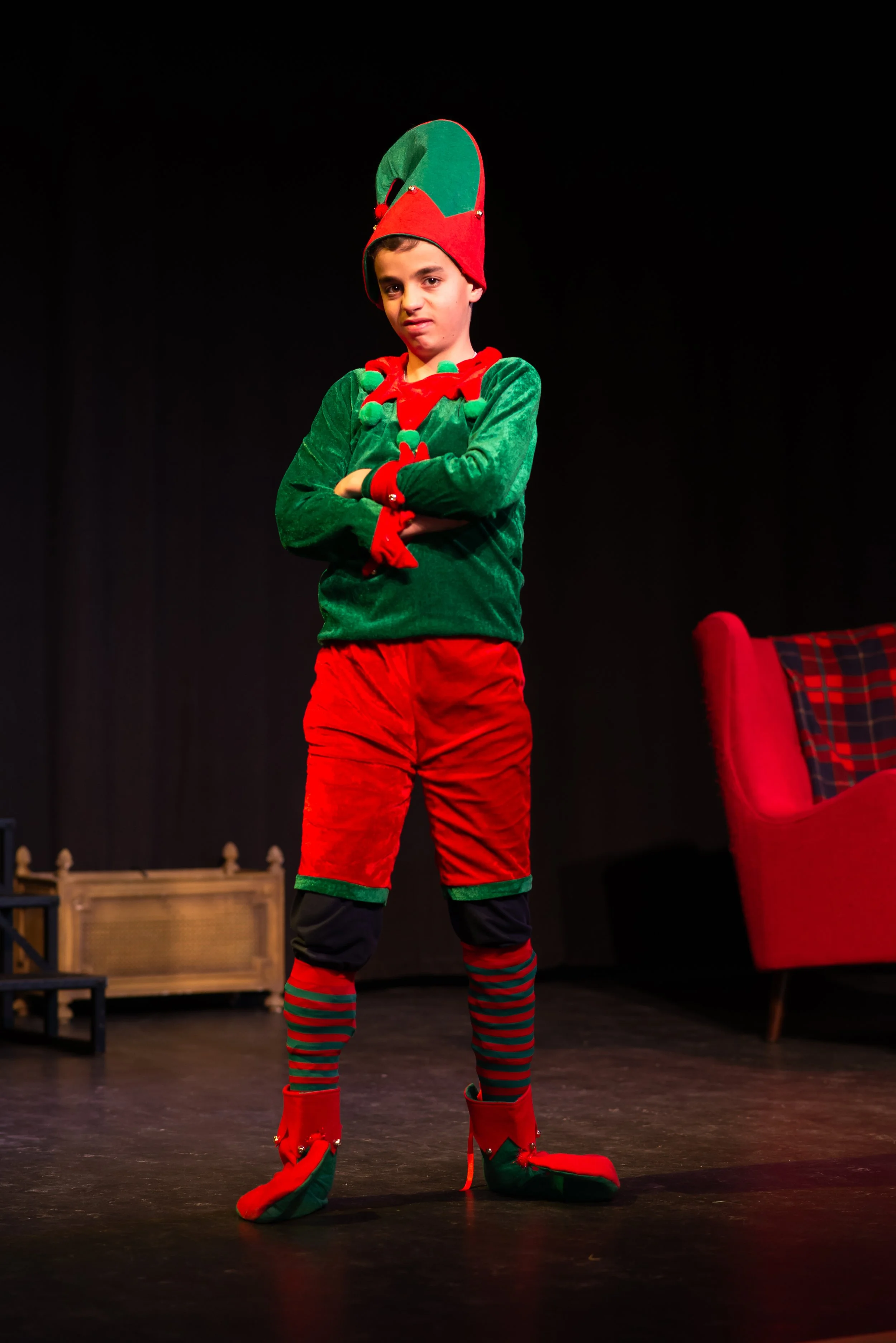 Young boy in an elf costume on stage with black background and red armchair nearby, arms crossed, wearing red and green striped stockings and elf slippers.