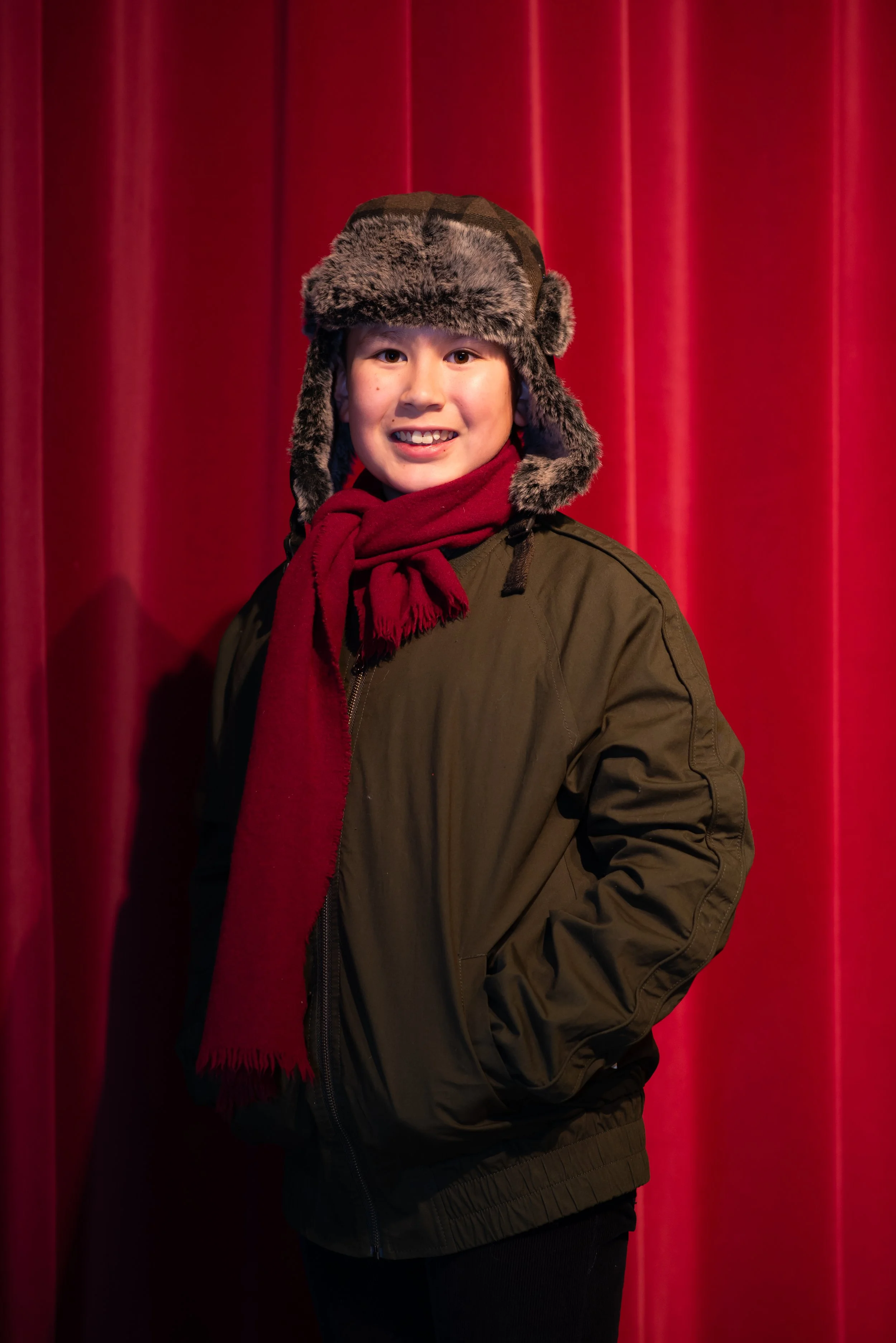 A young person smiling in front of a red curtain, wearing a furry winter hat, a red scarf, and a brown jacket.