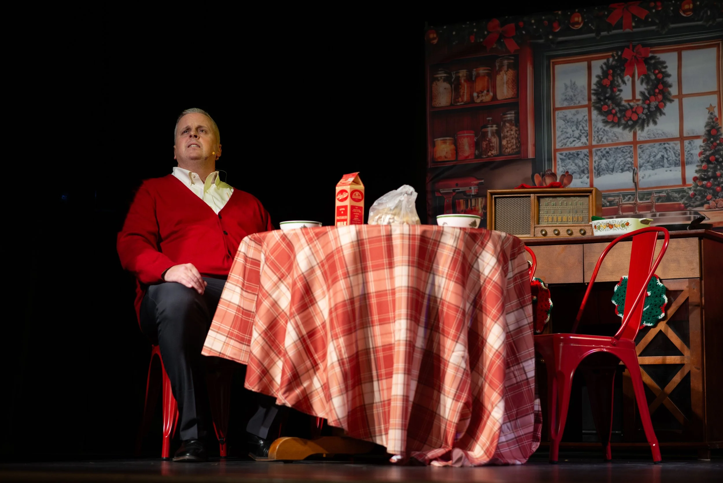 Man sitting at a holiday-themed table with a black background and a decorated window mural in the background.