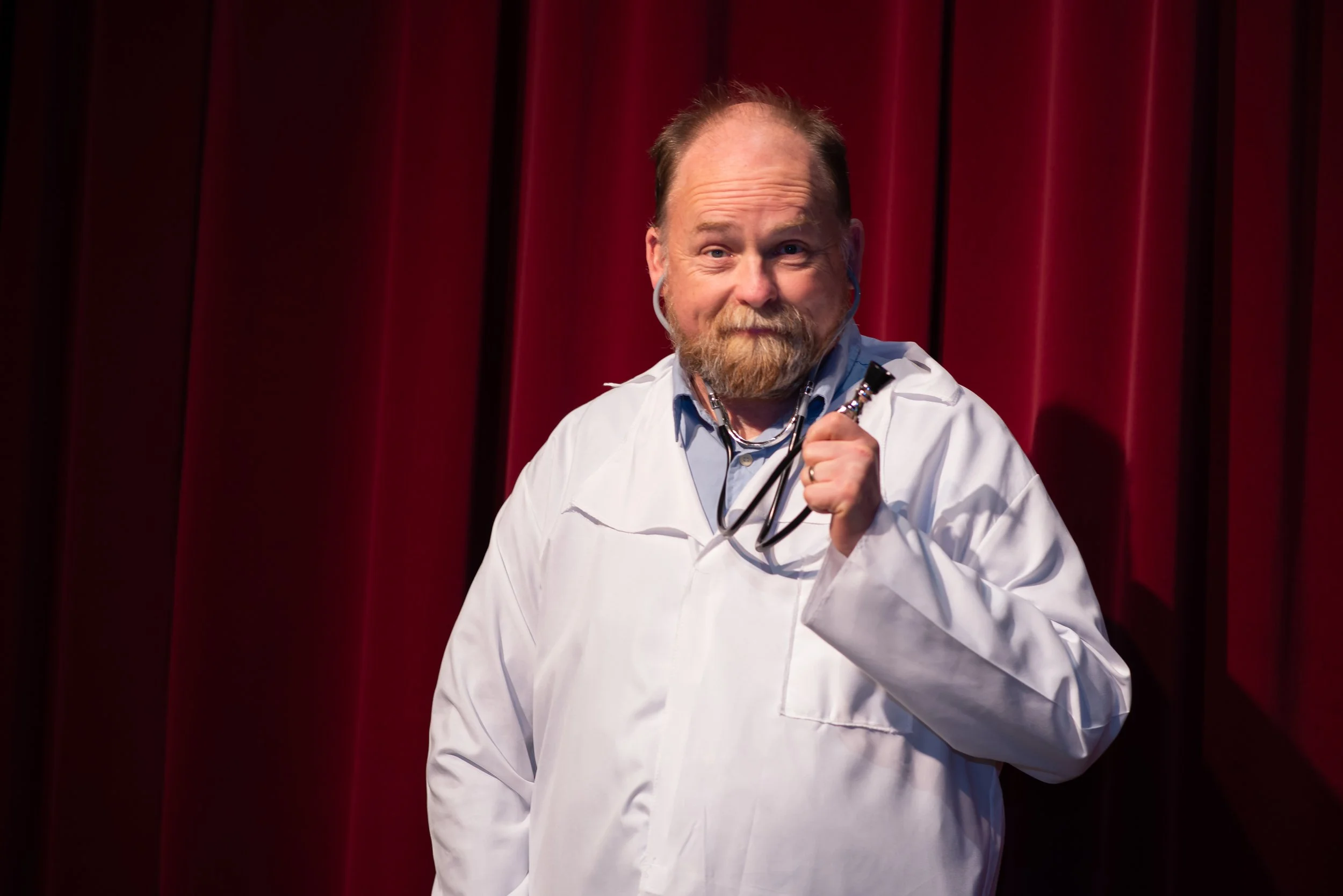 Man in a white lab coat holding a stethoscope, standing in front of a red curtain.