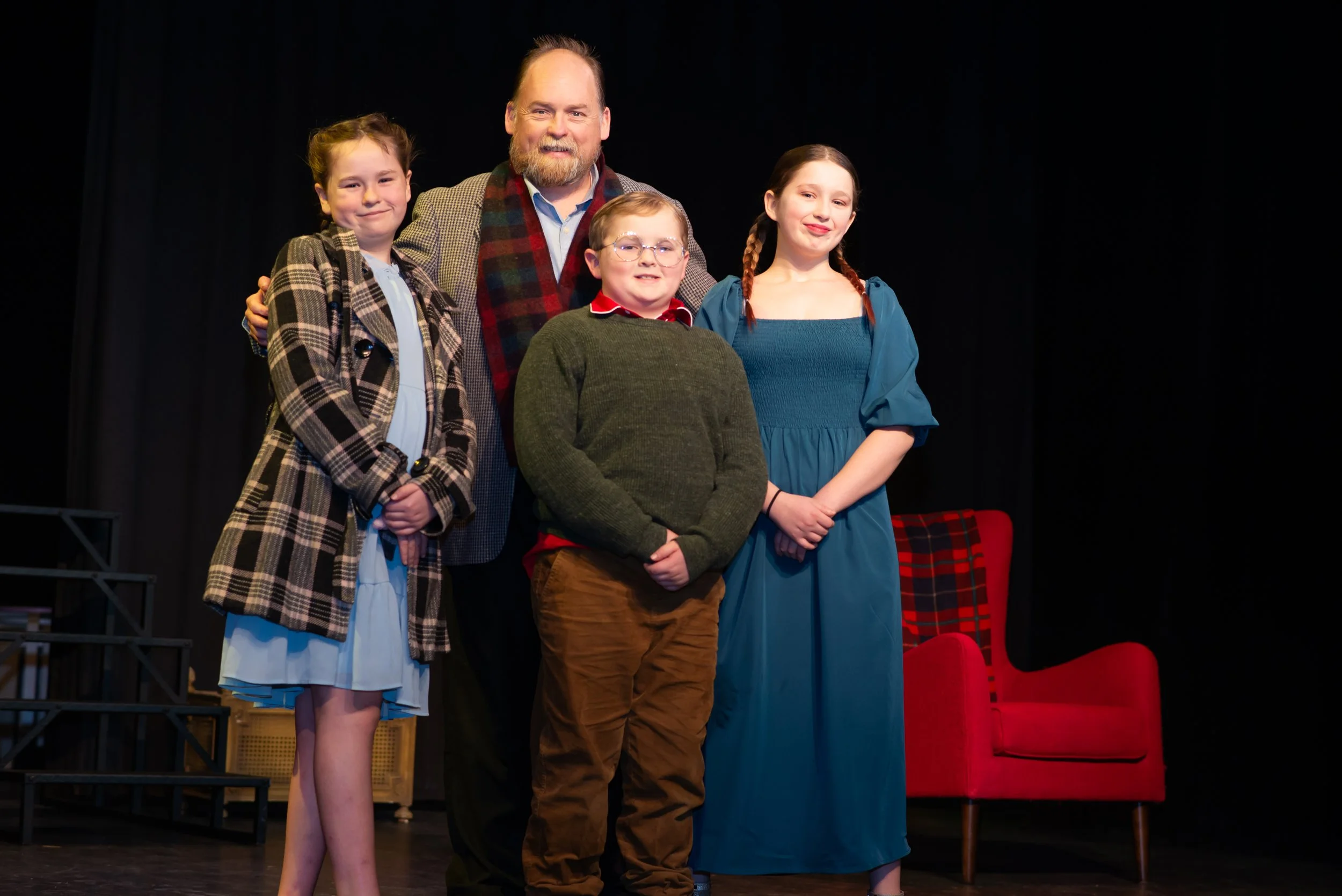 A group of five people, four children and one adult man, standing on a stage with a black background. The children are dressed in costumes, and the man is smiling, wearing a blazer and a plaid scarf. There are red chairs and stage equipment visible i