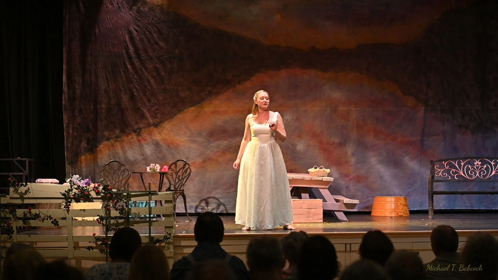 Female Jesus in a white dress standing on stage with a painted backdrop, props and chairs around, performing for an audience.