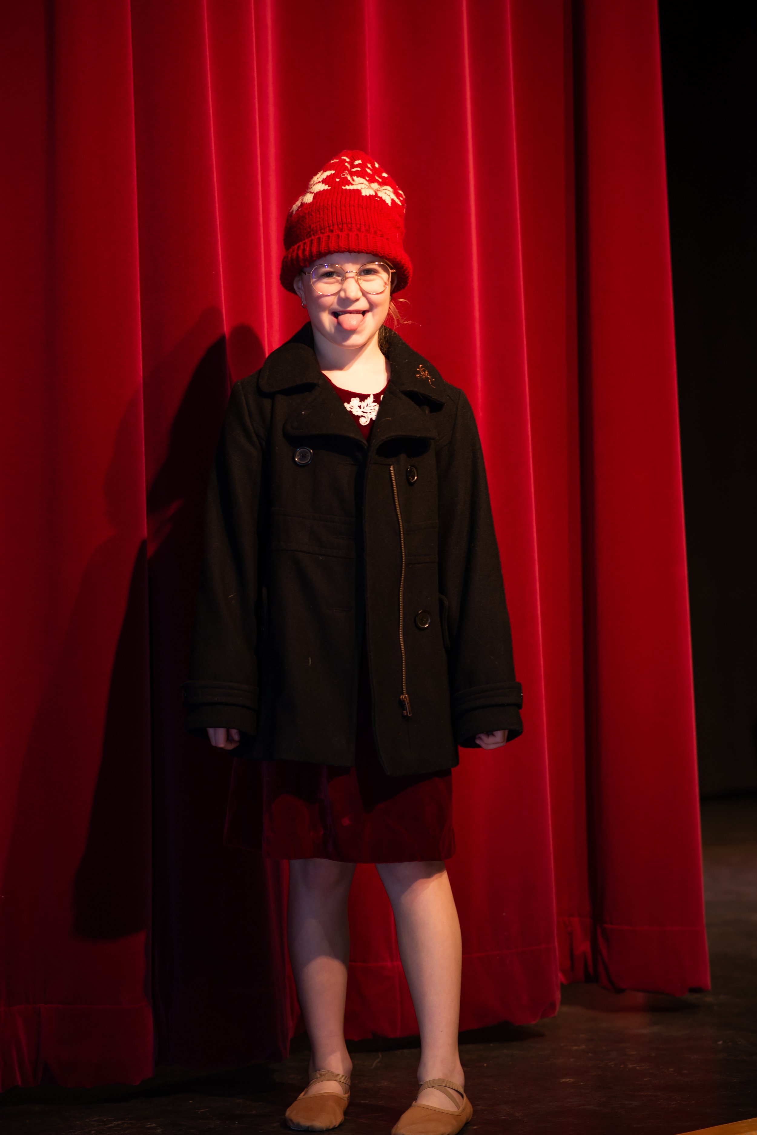 A young girl with glasses and a red knit hat with white patterns is standing in front of red stage curtains, sticking her tongue out and smiling.