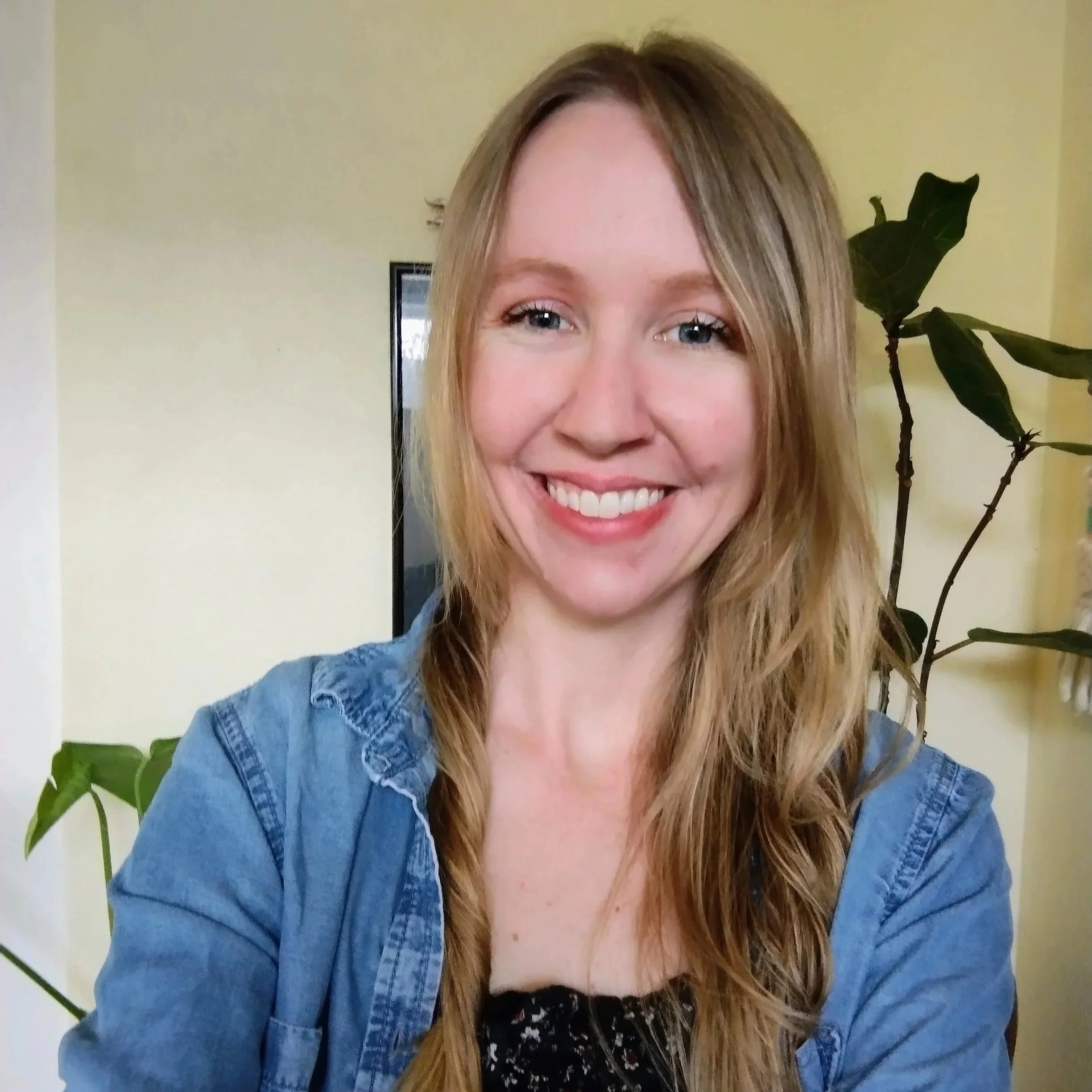 A woman with long blonde hair smiling at the camera, wearing a denim shirt, in front of a yellow wall and green plants.