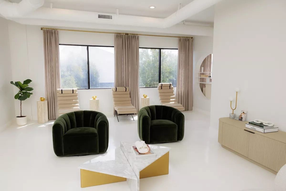Modern living room with white walls and floor, two dark green velvet armchairs, three beige lounge chairs placed near large front windows, a white marble and gold coffee table, a beige sideboard with books and a lamp, a potted plant, and beige curtains.