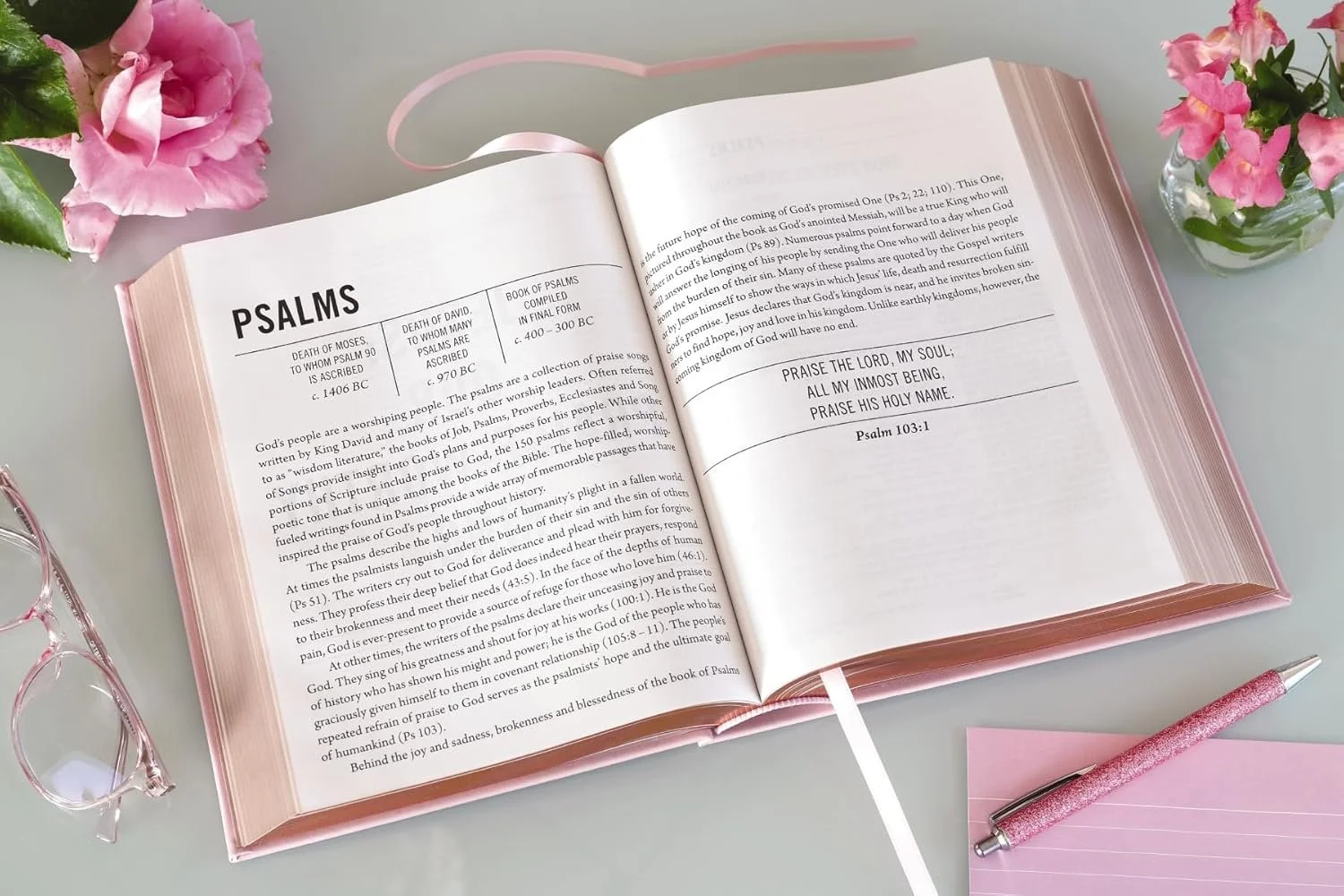 Open pink-bound book of Psalms with highlighted text, surrounded by pink roses, pink reading notepad, pink glitter pen, and pink glasses on a white surface.