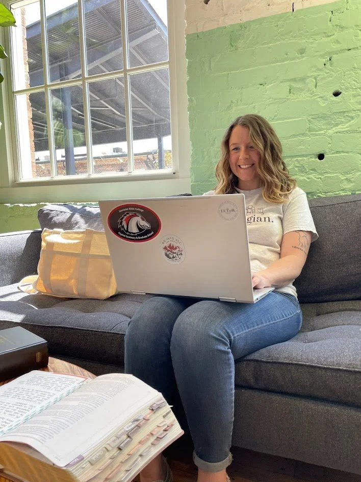 A woman sitting on a grey sofa working on a laptop, with a window behind her showing an outdoor building structure. The woman has curly blonde hair, is smiling, and is wearing a white t-shirt and blue jeans. There are books and a tote bag on the sofa and a large open book on a table in front of her.