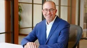 A smiling man in a blue blazer sitting at a desk in an office setting.