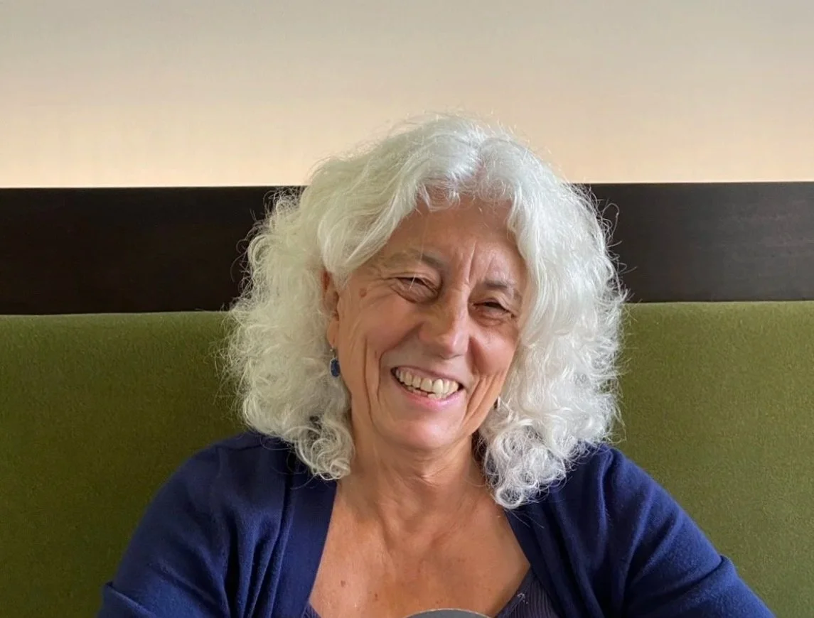 A woman smiling and wearing colorful glasses with cartoon characters, standing in a room with floral wallpaper, wooden cabinet, and a chair in the background.