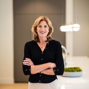 A woman standing indoors with her arms crossed, smiling, in front of a neutral background with modern lighting.