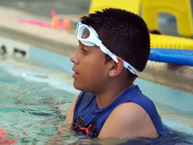 Boy with swim goggles resting at the edge of a swimming pool, wearing a blue swim shirt.