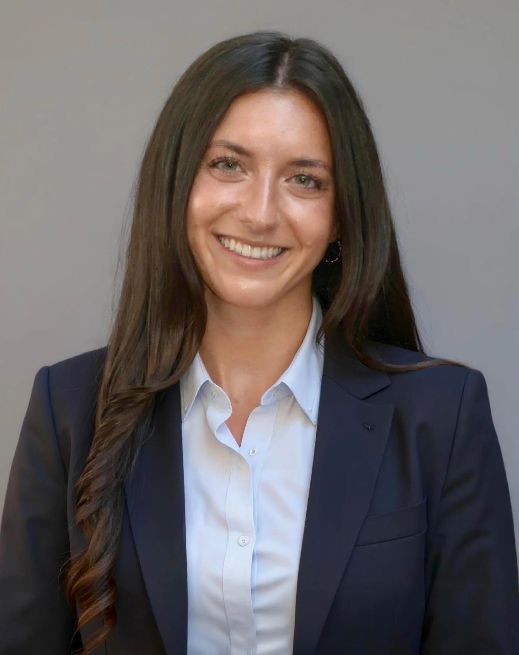 Professional woman with long dark hair, wearing a navy blazer and white shirt, smiling at the camera.