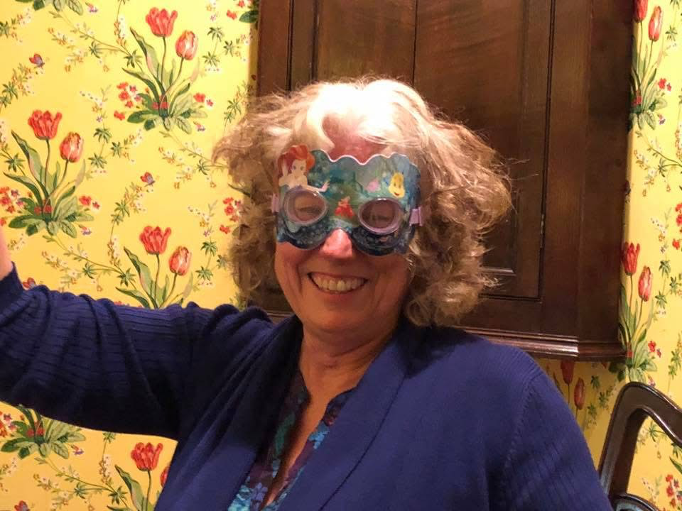 A woman smiling and wearing colorful glasses with cartoon characters, standing in a room with floral wallpaper, wooden cabinet, and a chair in the background.