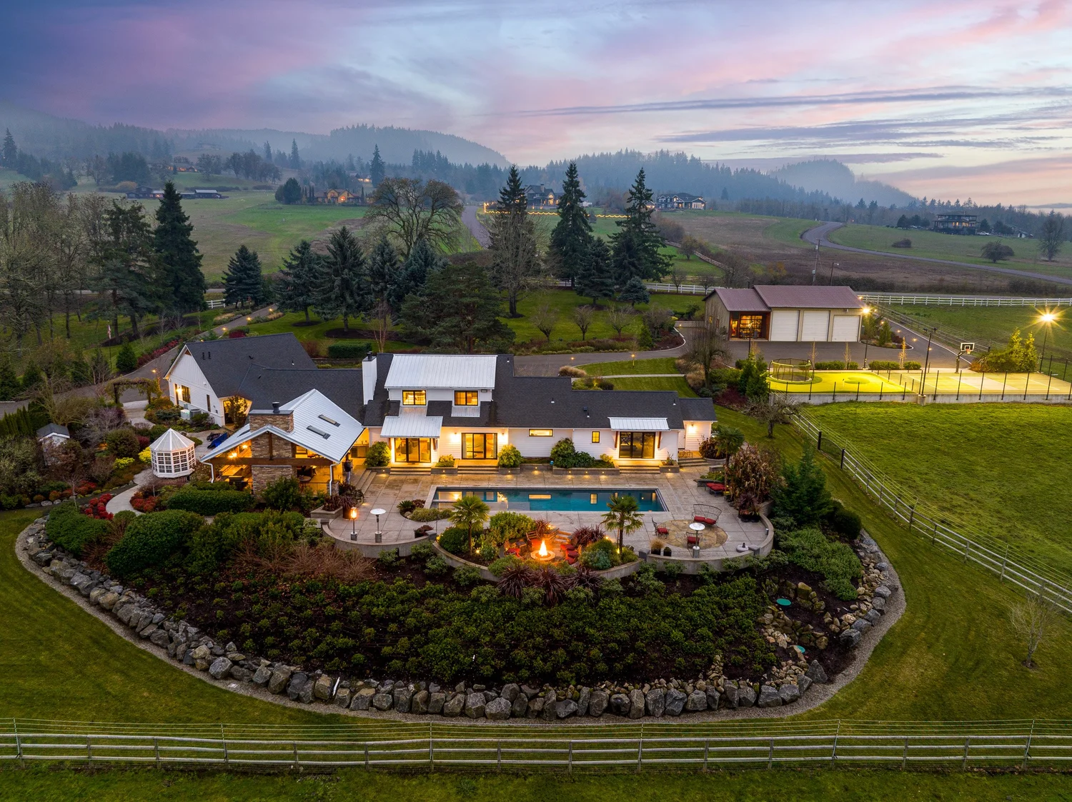 A spacious backyard with a pool, firepit, and landscaped garden overlooking rolling hills and trees at dusk