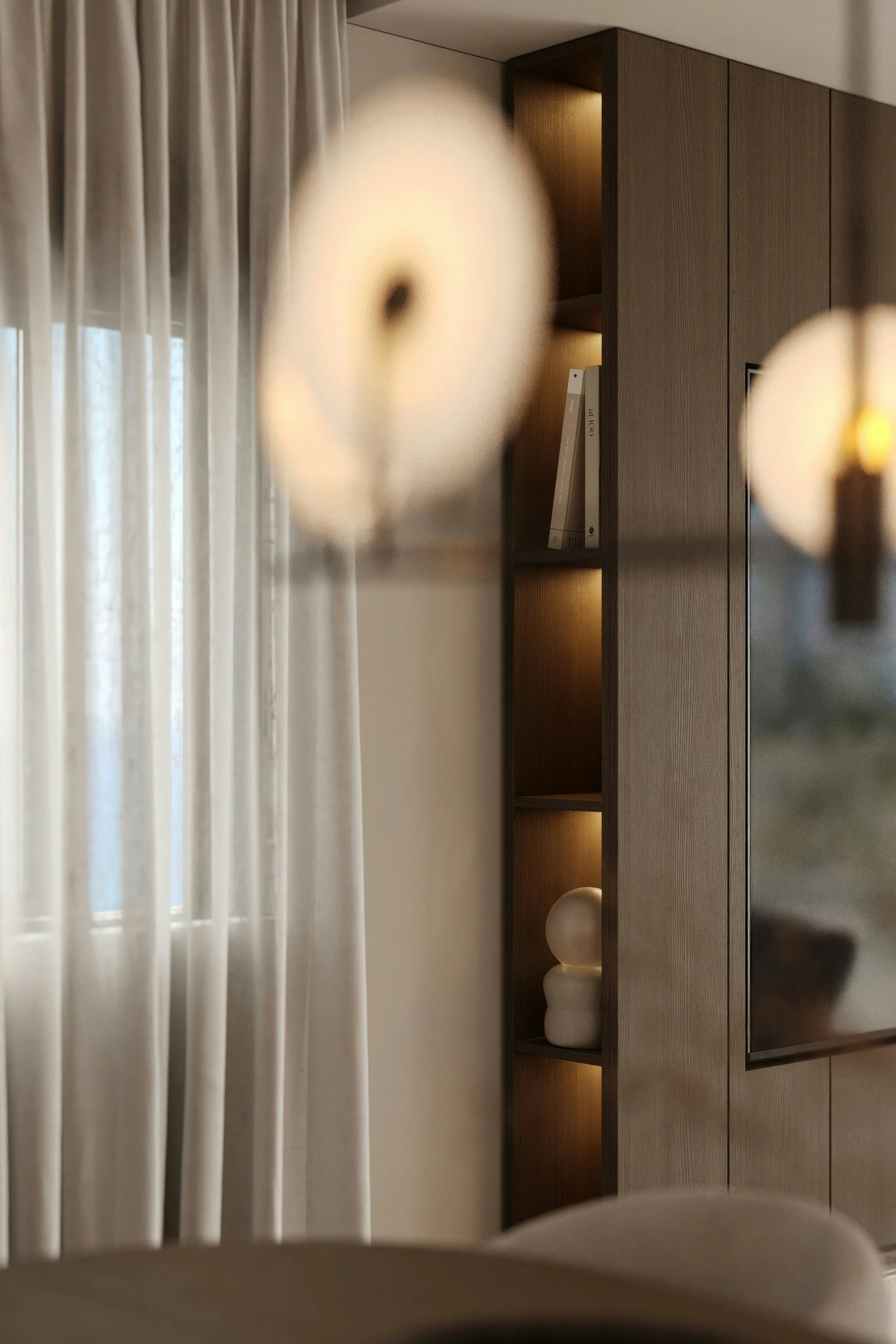 Close-up of two wall-mounted light fixtures in a modern living space, with a window with sheer curtains and a wooden shelf with decorative items in the background.