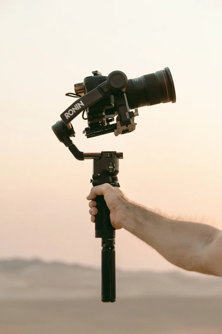 Cinematic real estate video production — videographer holding a camera mounted on a gimbal stabilizer against a blurred outdoor landscape at golden hour.