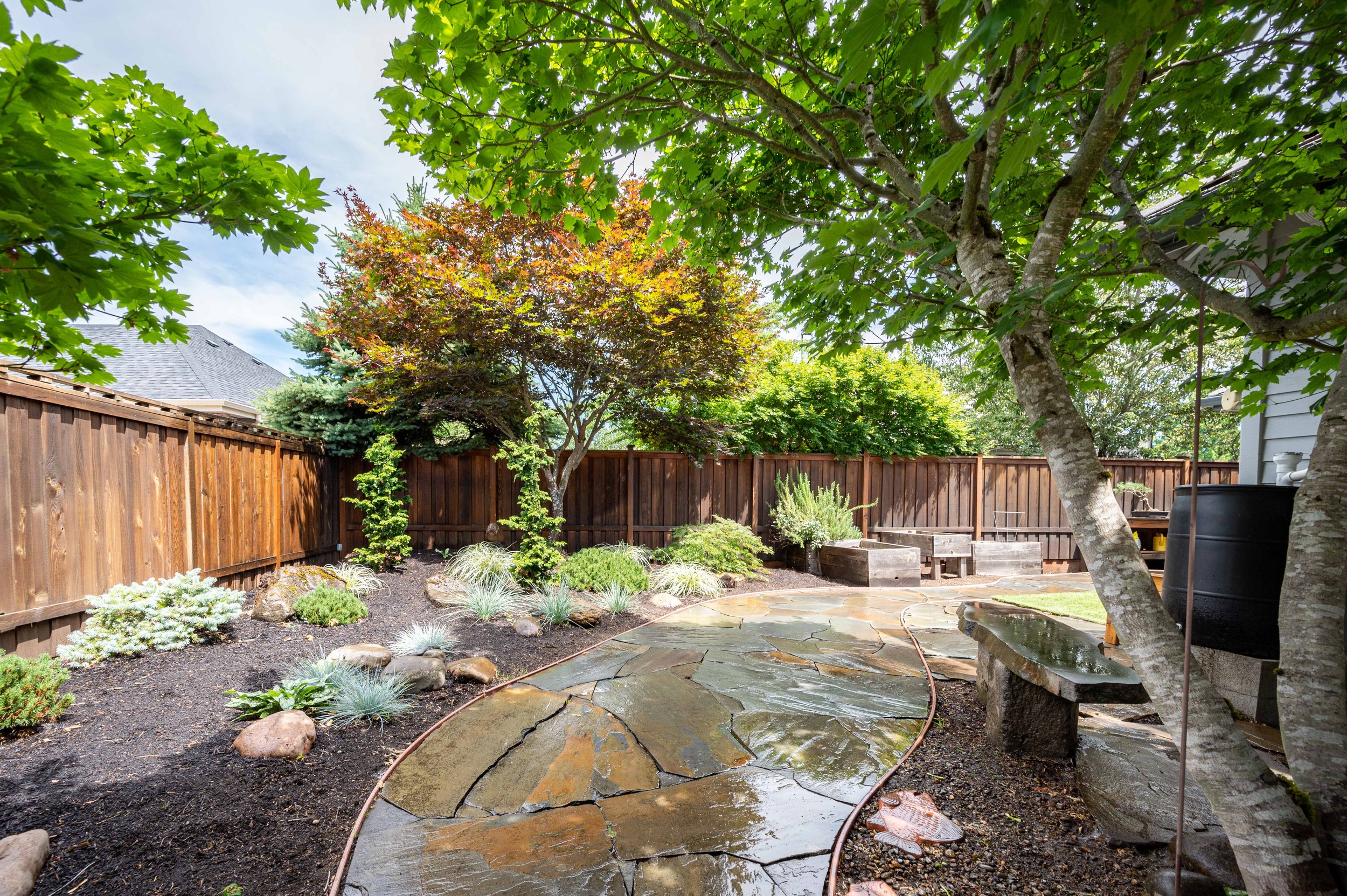 Backyard patio with stone walkway, mature trees, and landscaped garden beds