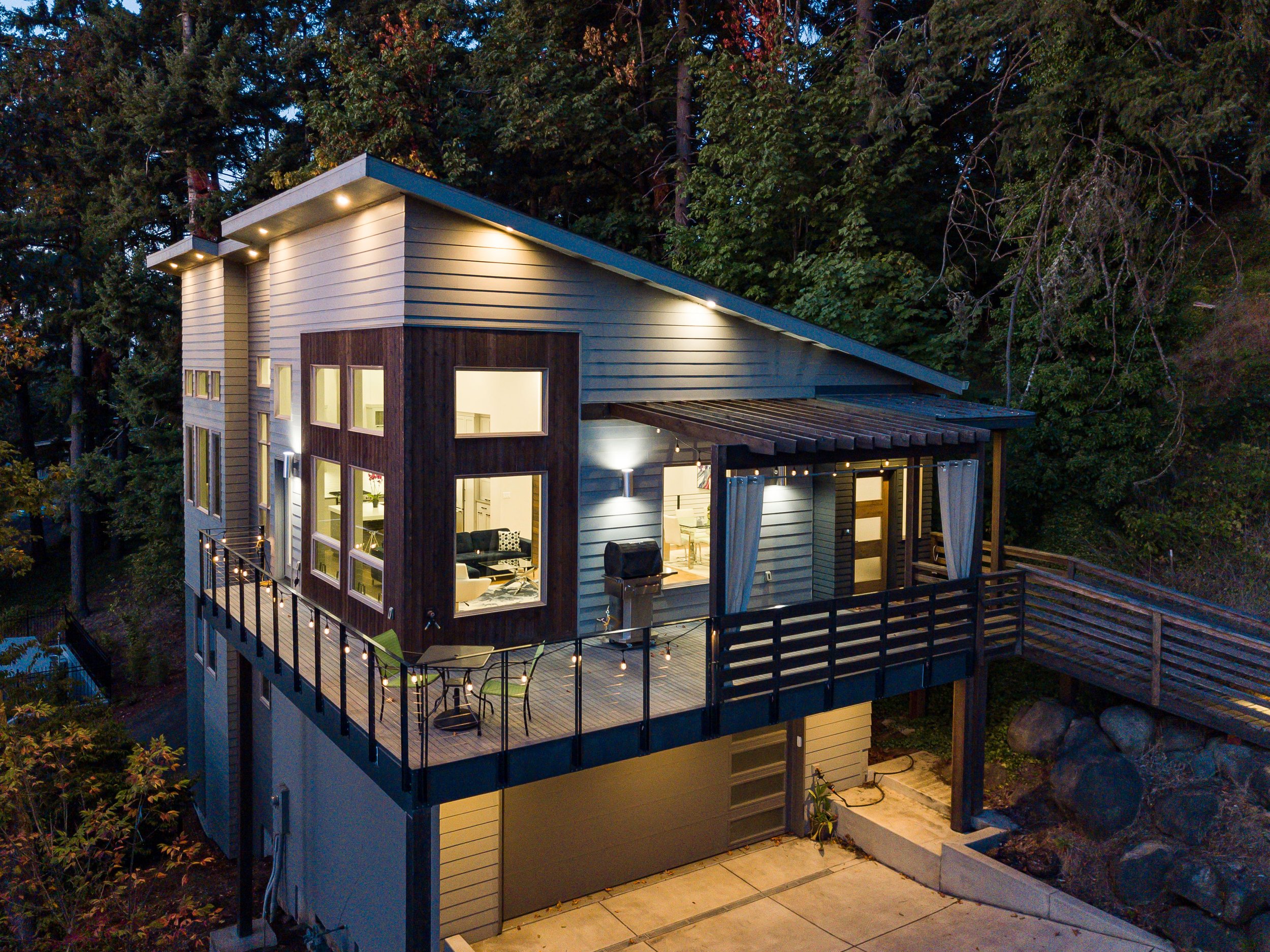 Modern two-story house with large windows, illuminated interior, outdoor deck with string lights, surrounded by trees at dusk.
