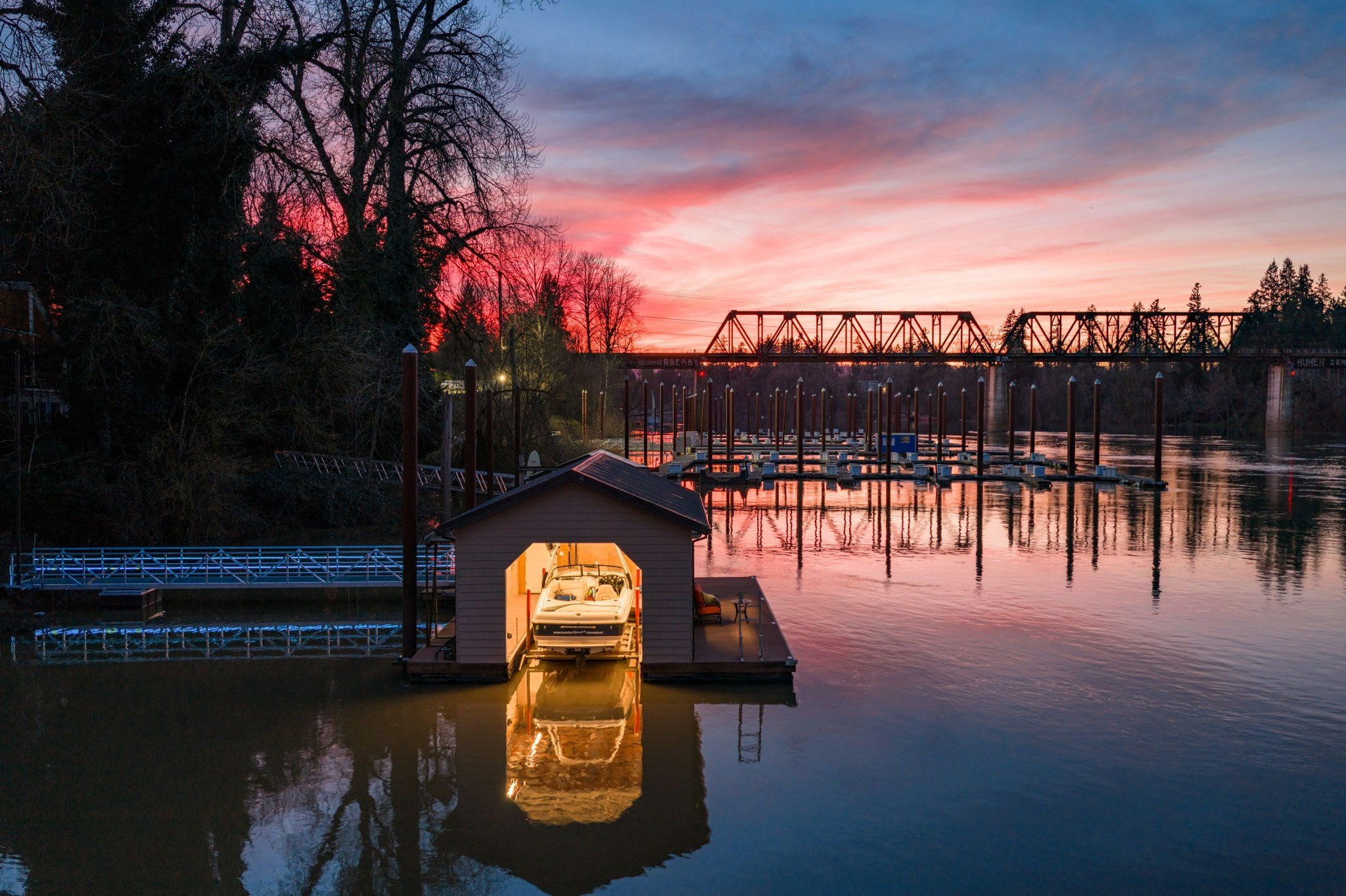 A boat dock on a river at sunset with a pink and blue sky, a boathouse with a boat inside, and a bridge in the background.