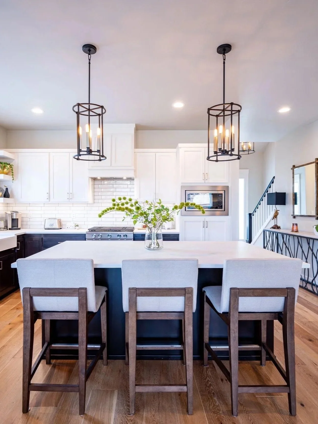 Bright modern kitchen with white shaker cabinets, subway tile backsplash, large island with three upholstered barstools, black metal pendant lights, and warm hardwood floors in a Portland home.