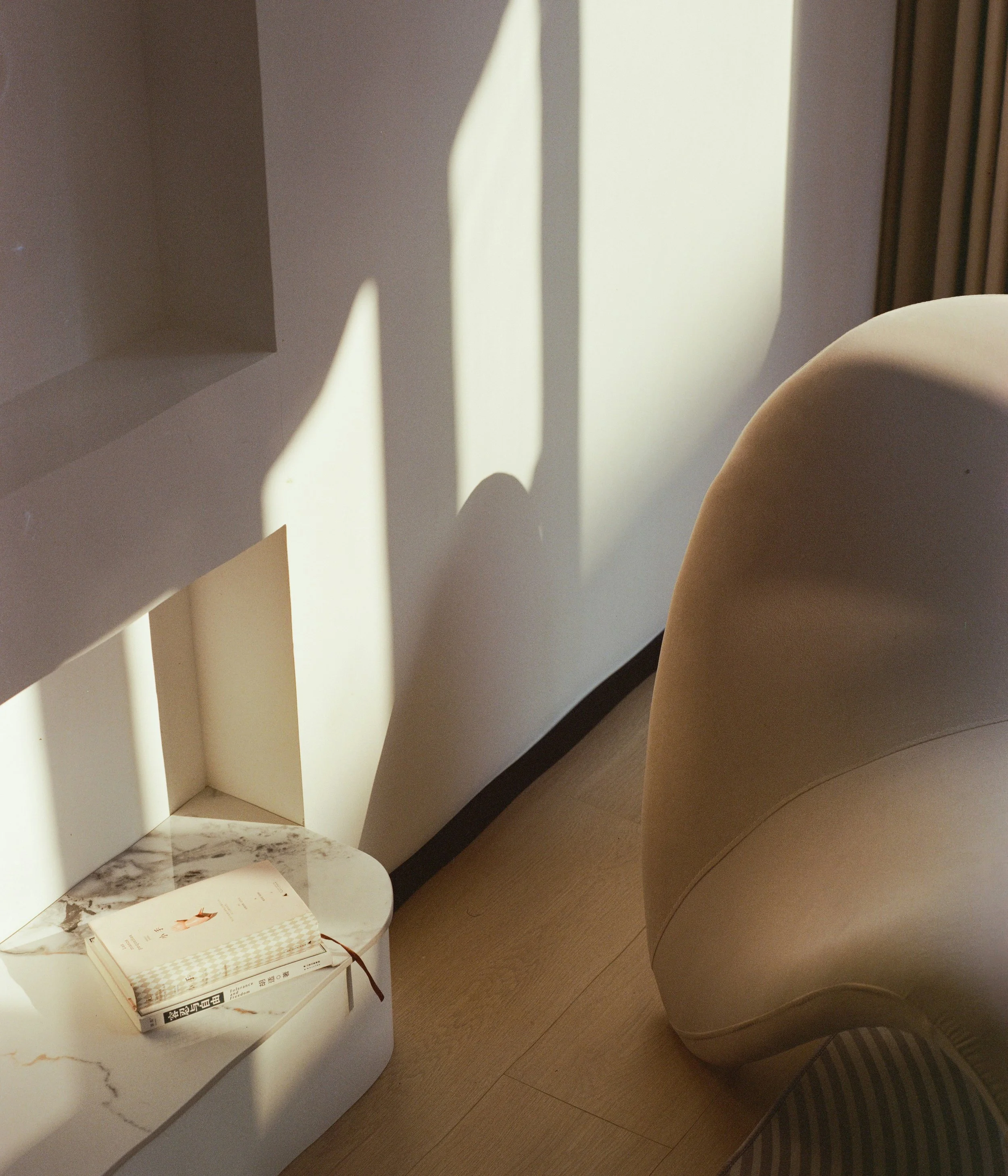 A corner of a room with sunlight casting geometric shadows on a white wall. There is a white, circular side table with a closed book on it, next to a beige, rounded chair on a wooden floor.