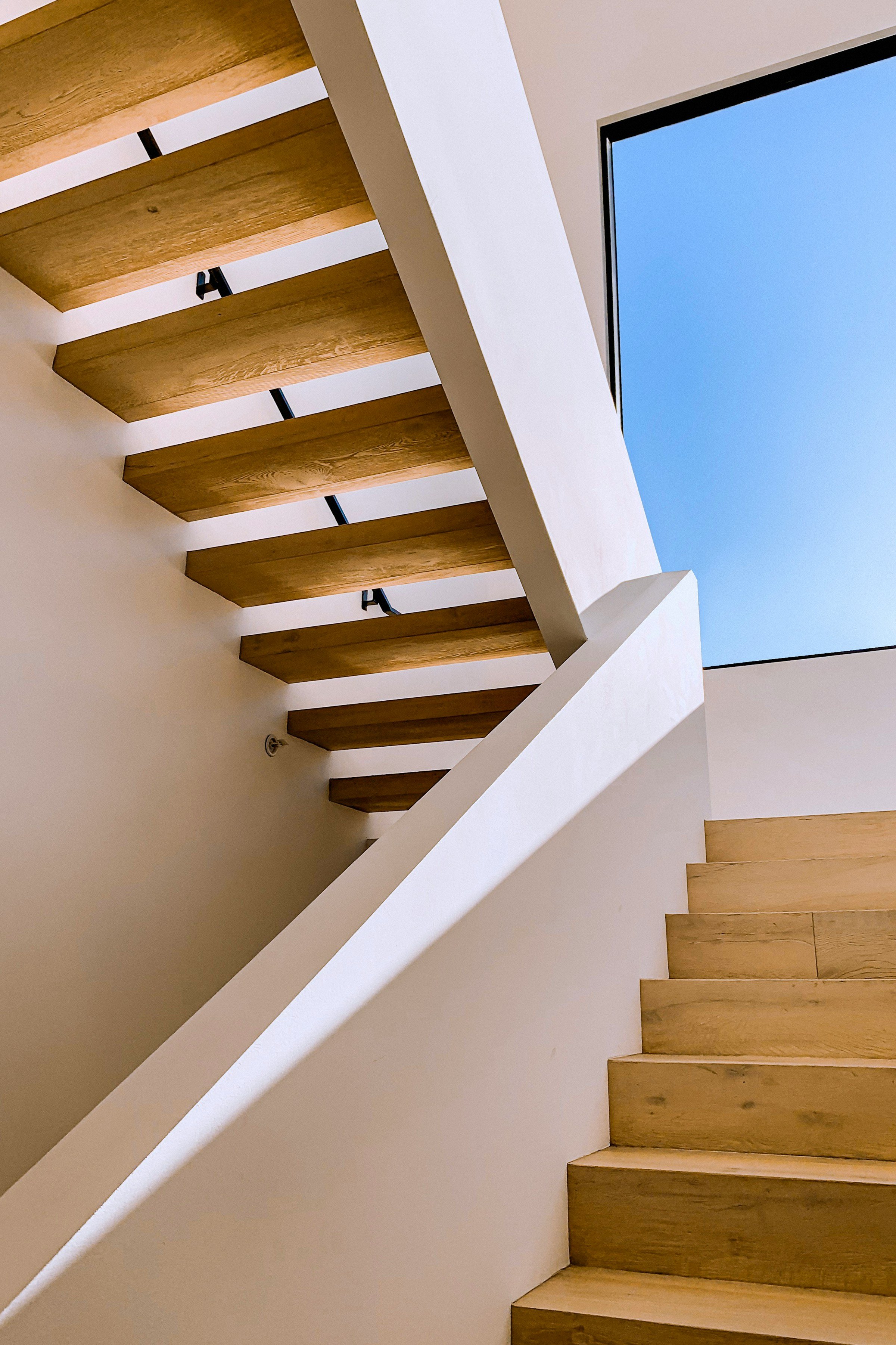 Modern floating wood staircase with white walls and skylight bringing in natural light against a clear blue sky.
