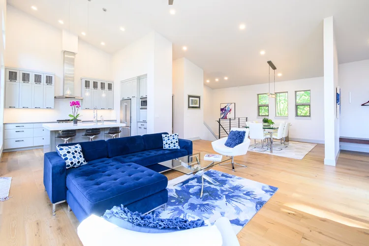 Real estate photography — bright open-concept living room and kitchen with modern white cabinets, stainless steel appliances, blue sofa, white chairs, and a blue and white area rug.