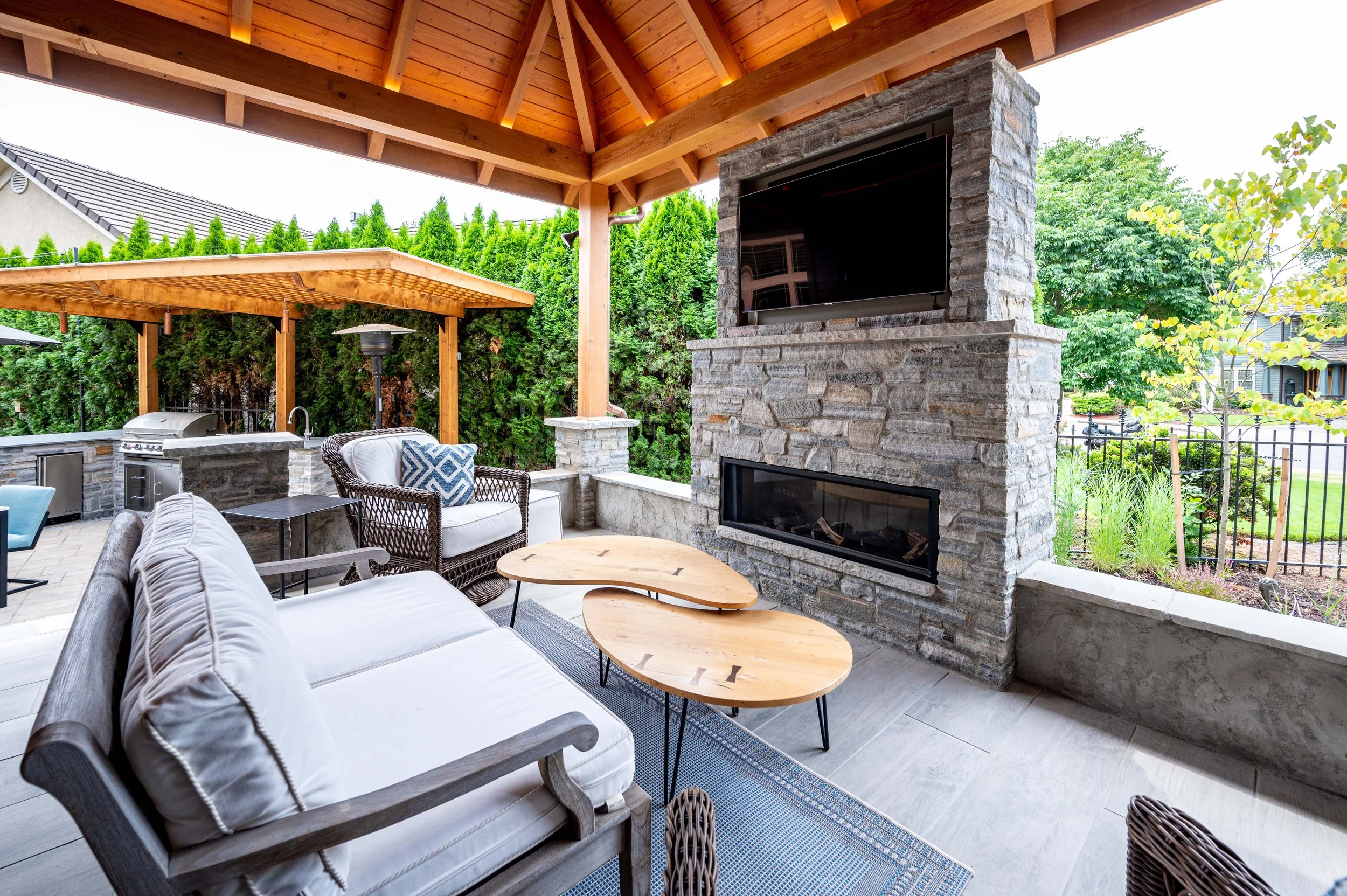 Covered outdoor living area with stone fireplace, mounted TV, seating, and wood pergola