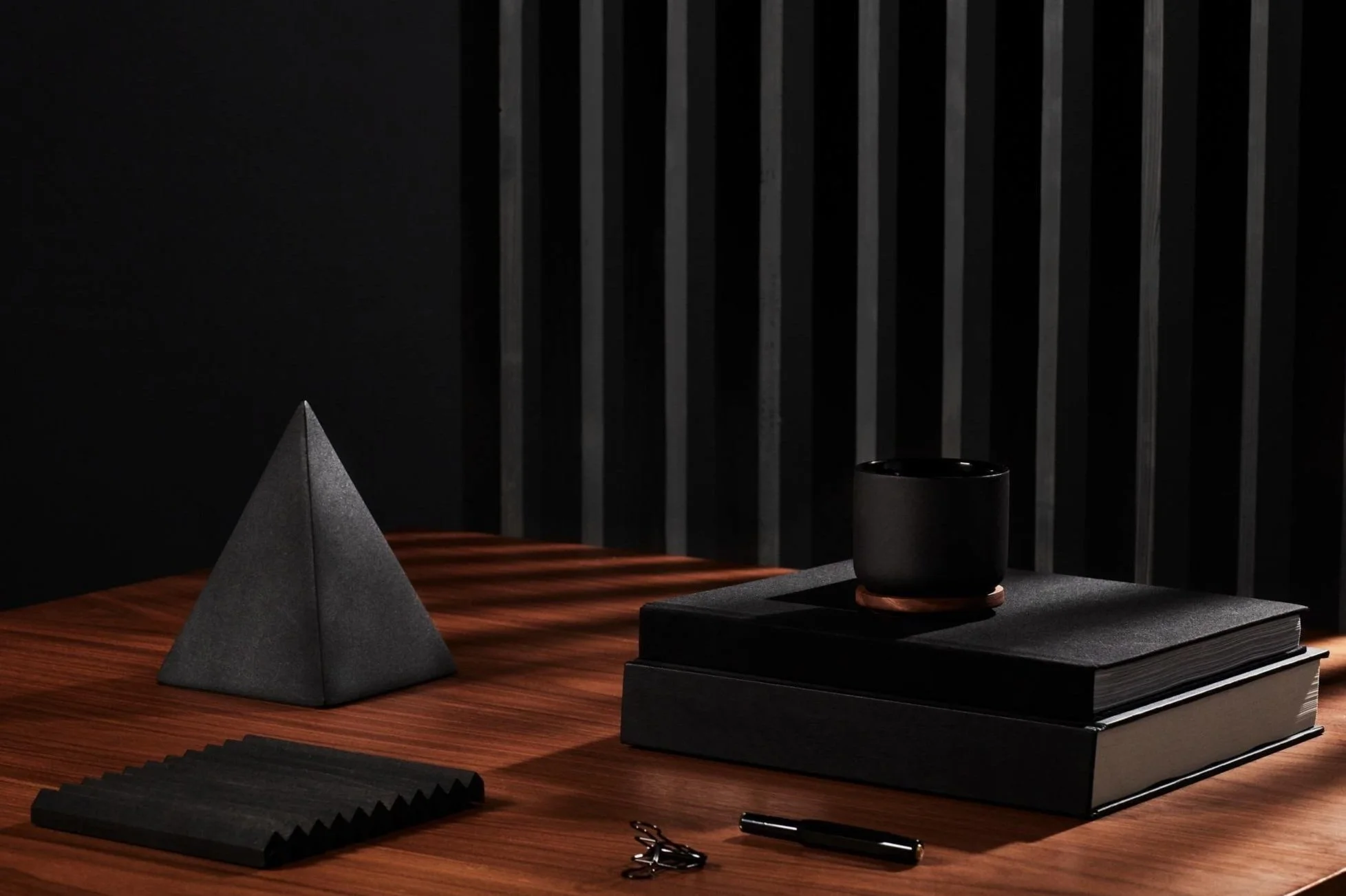A dark office desk scene with a black pyramid-shaped decor, black notebooks stacked, a black cup on a coaster, a set of black notecards, a pair of scissors, and a black pen, with vertical black slats in the background.