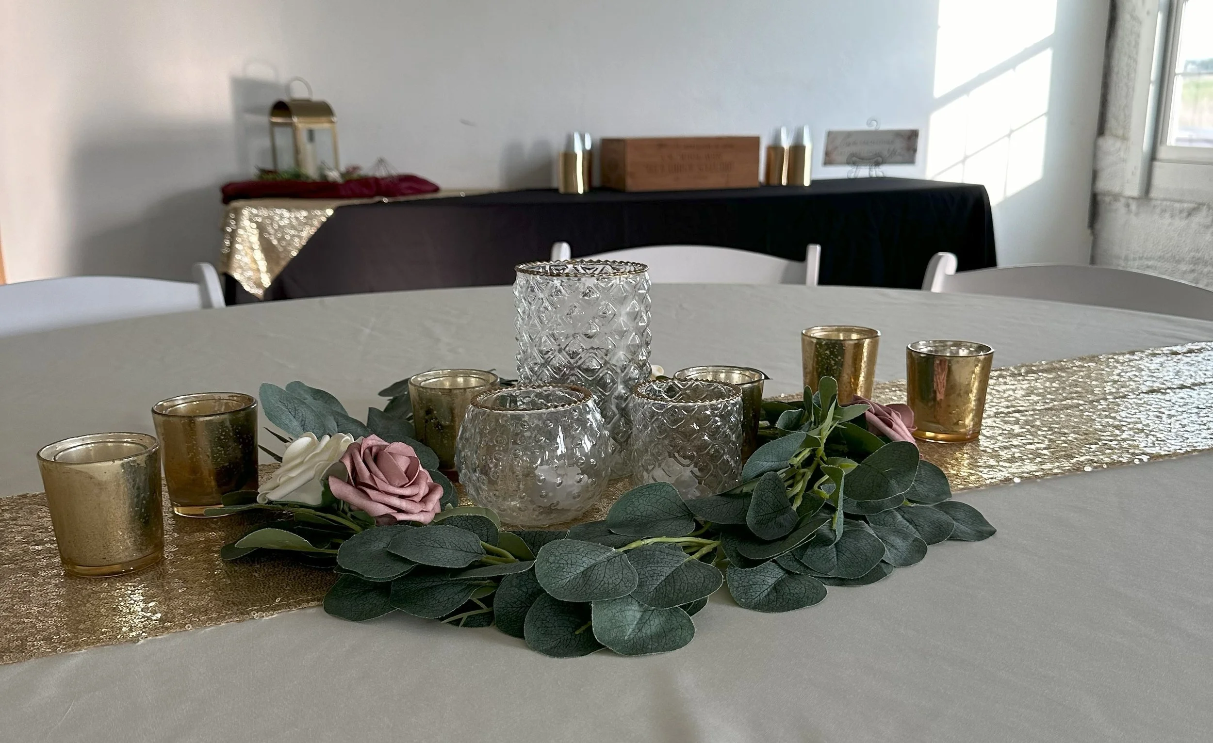 This is a beautiful table centerpiece for a wedding. I uses gold-rimmed glass vases, gold votive candles, eucalyptus, and faux foam flowers in mauve, pink, and white. It looks stunning on the gold sequin runner.