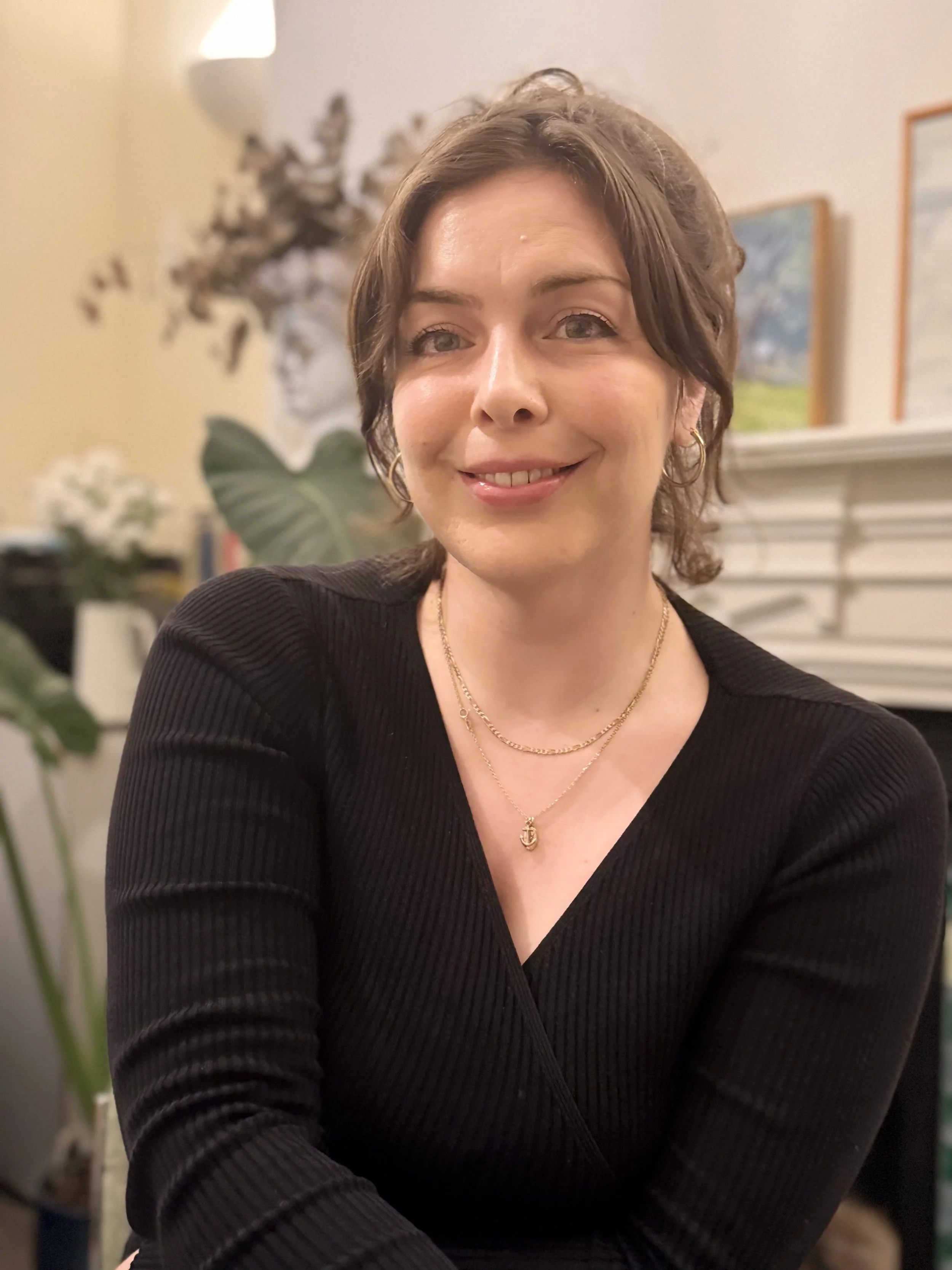 A woman with short brown hair and earrings, wearing a black V-neck sweater and gold jewelry, smiling in an indoor setting with plants and framed pictures in the background.
