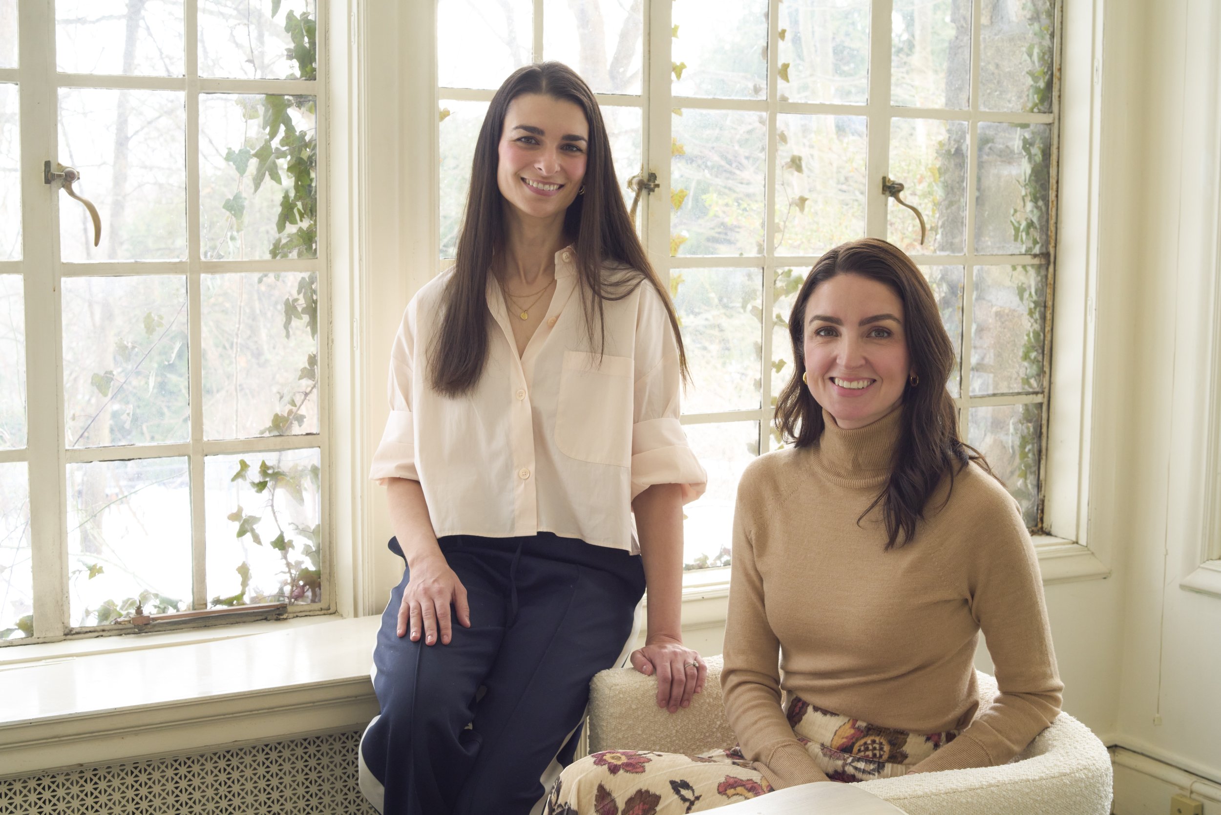 Two women smiling indoors near a large window. One woman is standing, wearing a light-colored blouse, while the other is seated, wearing a light brown turtleneck and floral skirt.