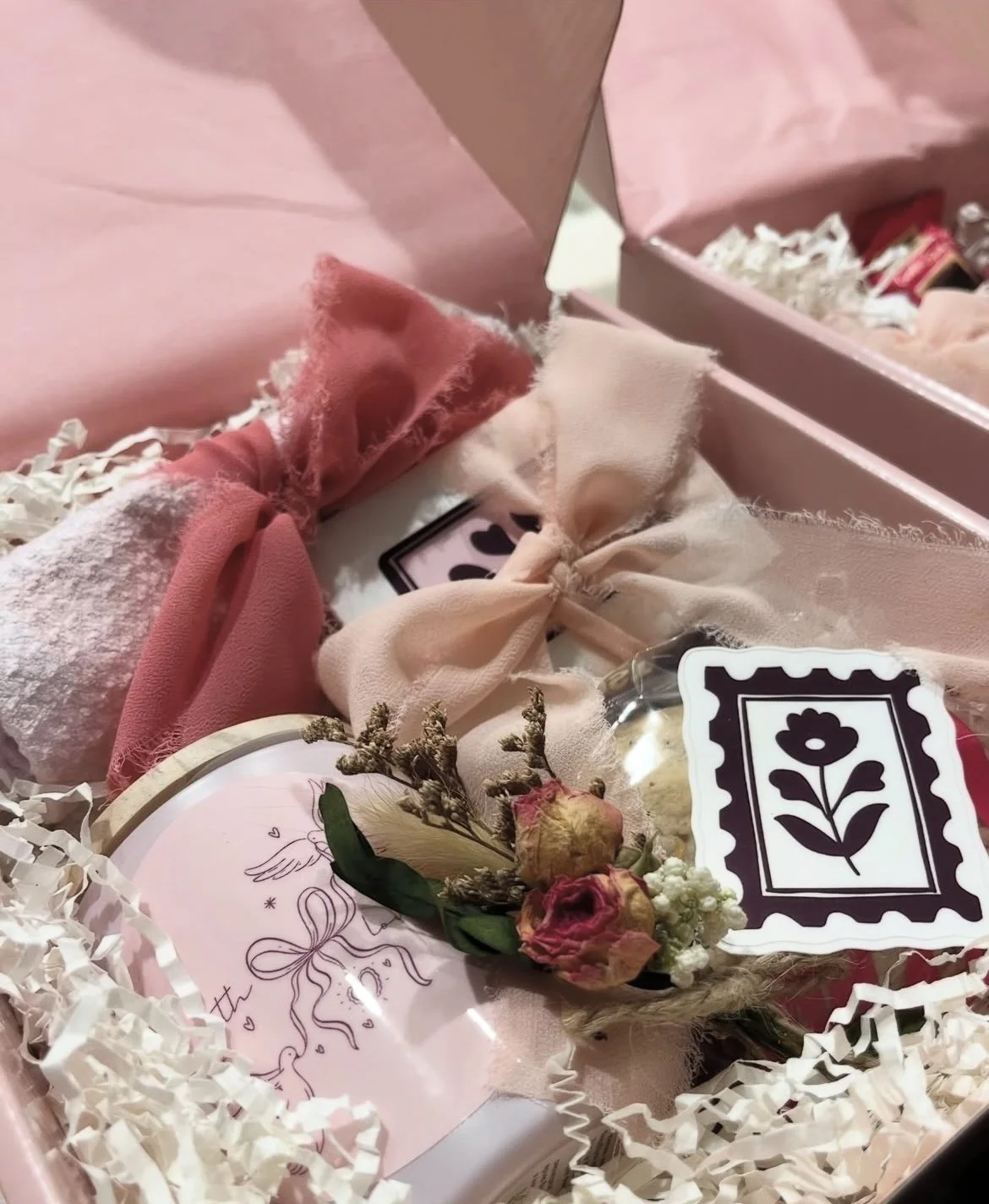 A pink gift box filled with tissue paper, a small bouquet of dried flowers, a pink card with a bow illustration, and decorative items wrapped in pink and white fabric, all arranged neatly.
