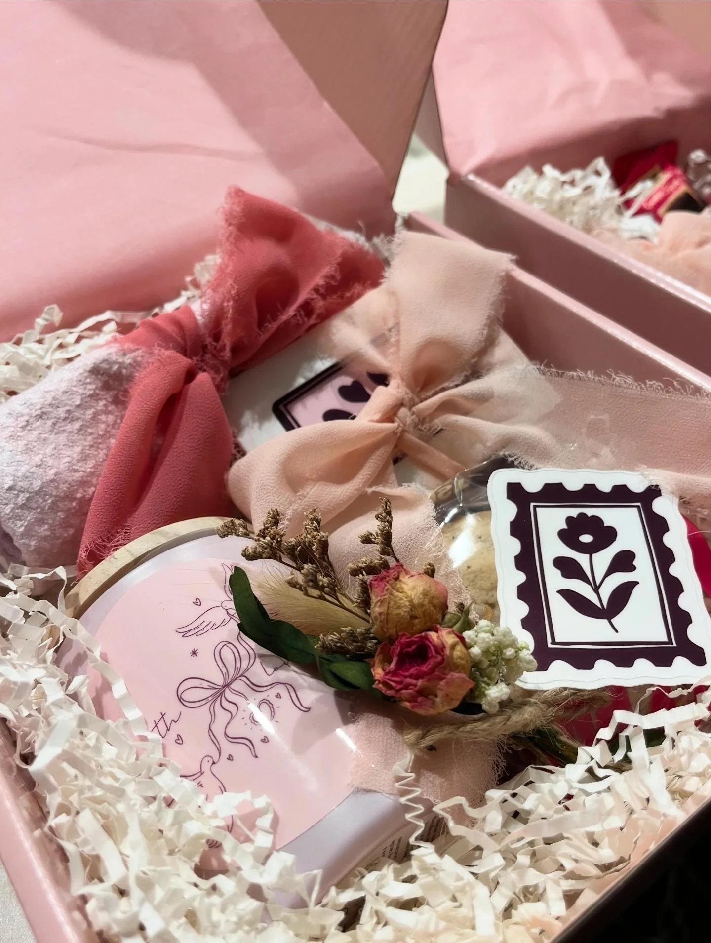 Pink gift box filled with shredded paper, containing assorted items such as a floral arrangement, a card with a unicorn, and other pink and neutral-colored accessories.